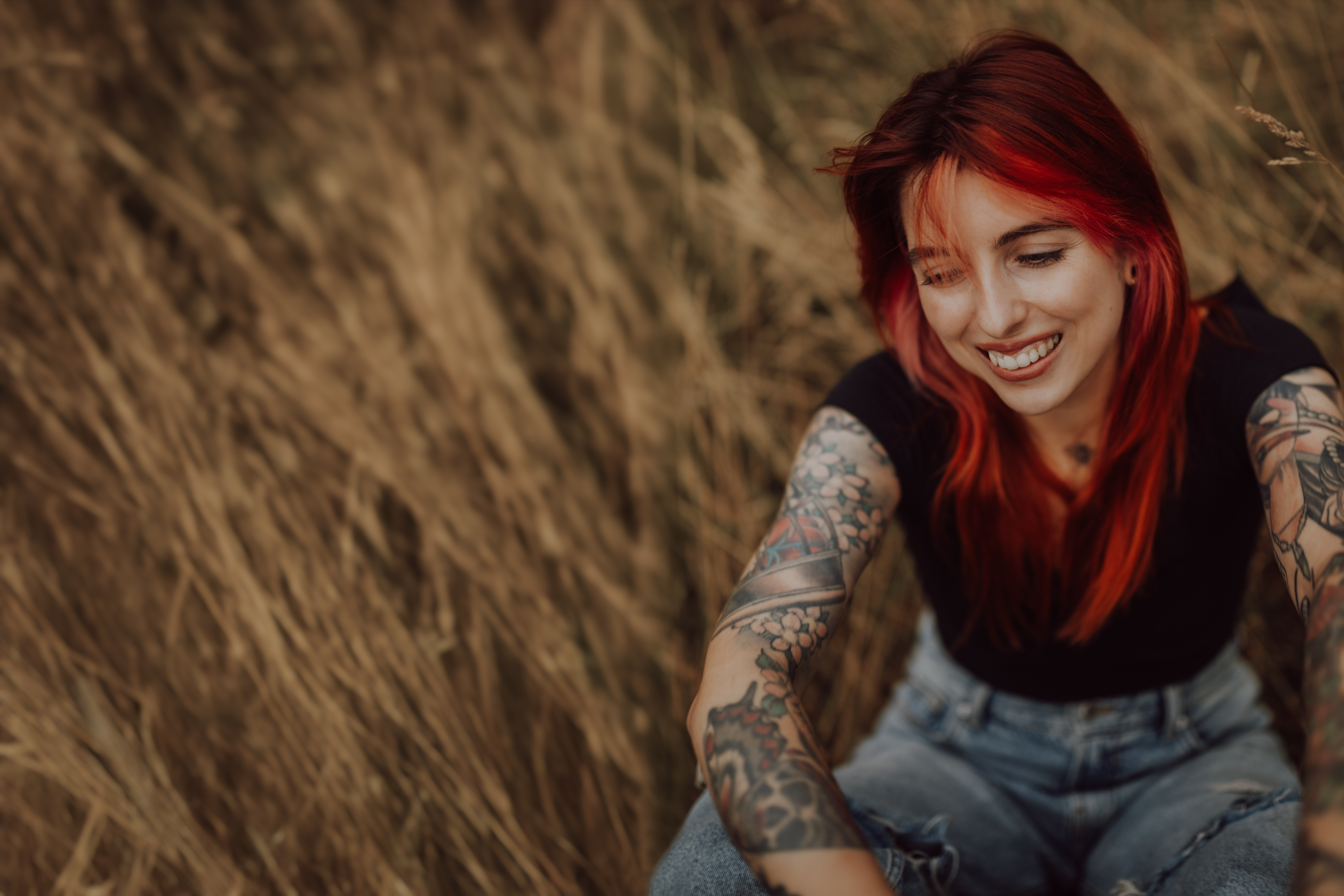 Eine Person mit leuchtend roten Haaren und Tattoos auf beiden Armen sitzt in einem Feld aus hohem, trockenem Gras. Sie trägt ein schwarzes Hemd und Jeans und lächelt, während sie während eines Fotoshootings im Aargau nach unten blickt.