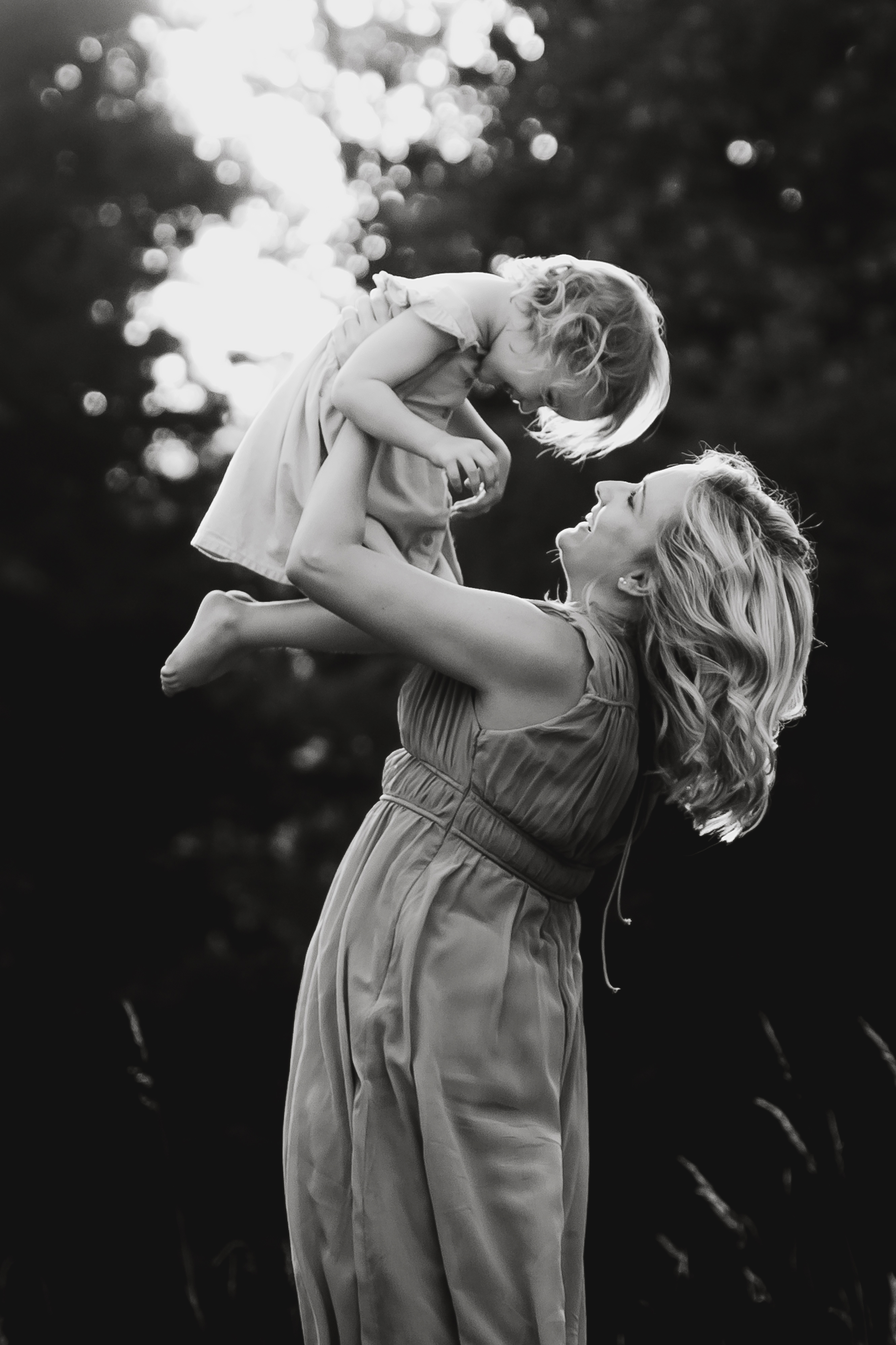 Ein Schwarzweißfoto von Tasha Ender, die ein kleines Kind über ihren Kopf hebt. Beide lächeln und schauen sich in die Augen, was einen Moment der Freude und Verbundenheit widerspiegelt. Der Hintergrund ist eine verschwommene Naturkulisse mit Bäumen, die die Essenz ihres Fotoshootings in Baden einfängt.