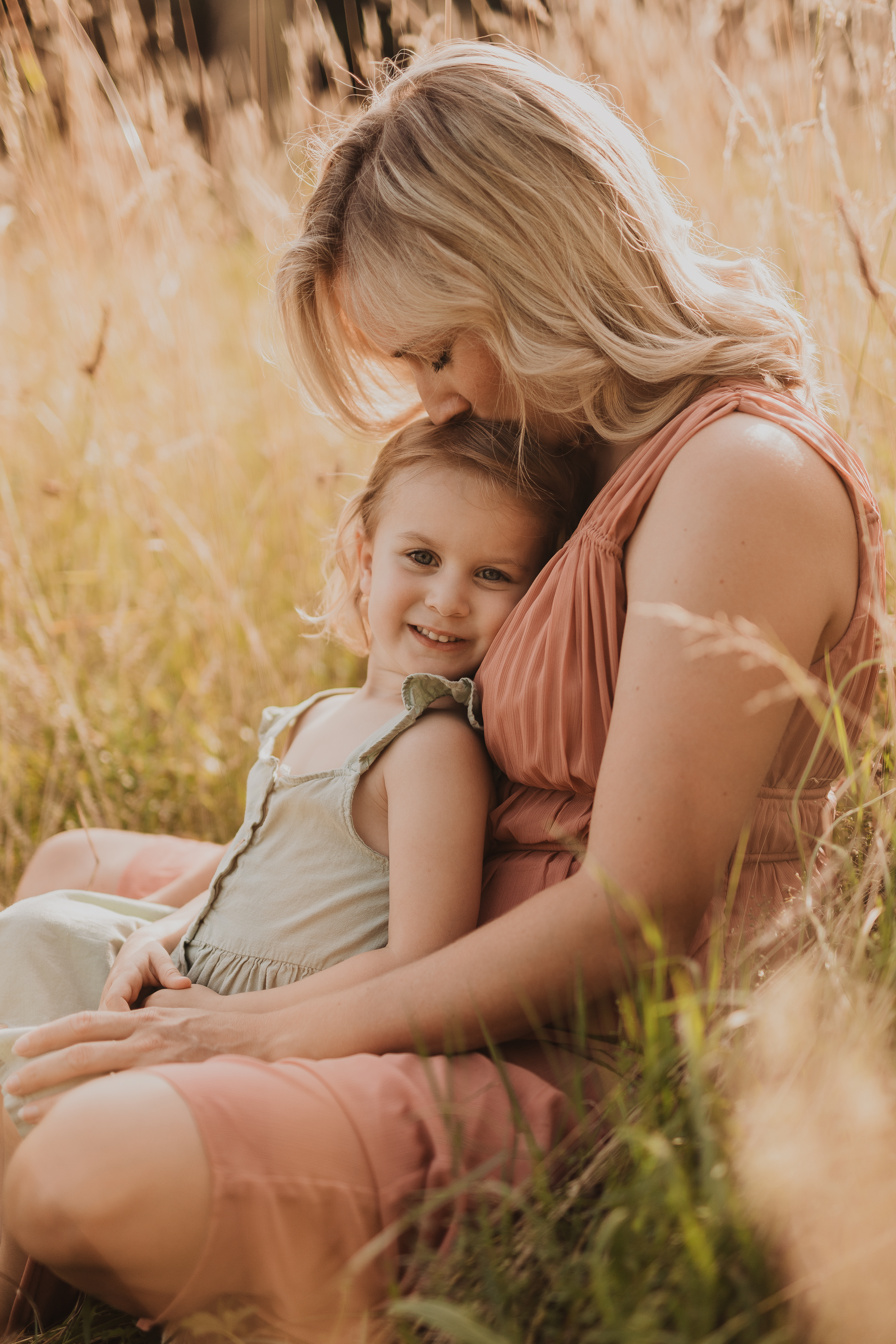 Eine Frau mit blondem Haar umarmt ein kleines Kind auf einem Feld mit hohem, goldenem Gras. Die Frau, die ein pfirsichfarbenes Kleid trägt, küsst liebevoll den Kopf des Kindes. Das Kind, lächelnd und in einem hellen Kleid gekleidet, sitzt während ihres Fotoshootings mit Tasha Ender in Baden zufrieden auf dem Schoß der Frau.