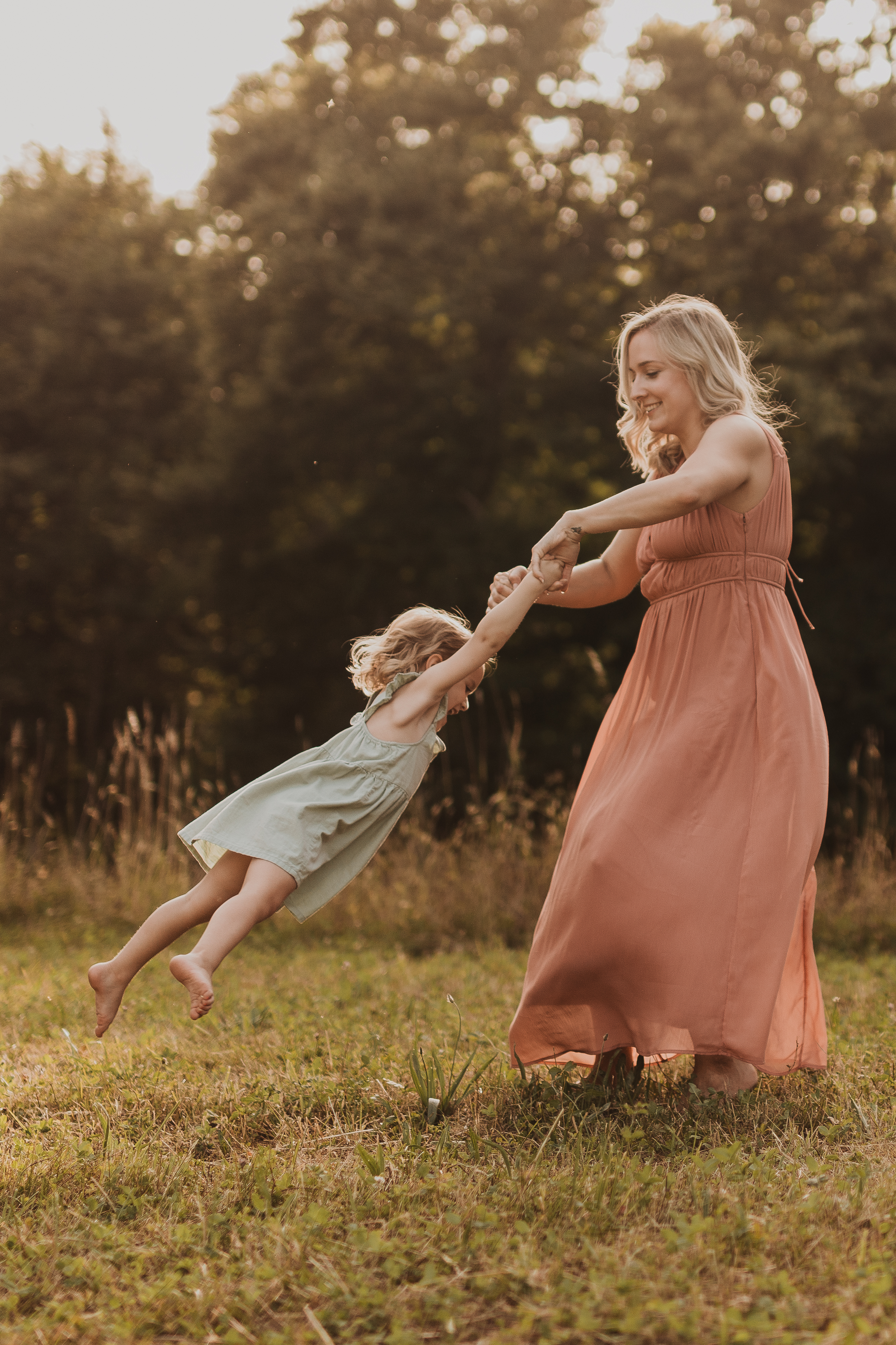 Eine Frau in einem langen, pfirsichfarbenen Kleid dreht ein kleines Kind in einem hellgrünen Kleid auf einer Wiese. Das Kind lacht, als es vom Boden hochgehoben wird, während die Frau lächelt und diesen Moment für natürliche Familienfotos festhält. Bäume und Büsche bilden eine üppige, grüne Kulisse, die perfekt für jede Fotografin in Baden ist.