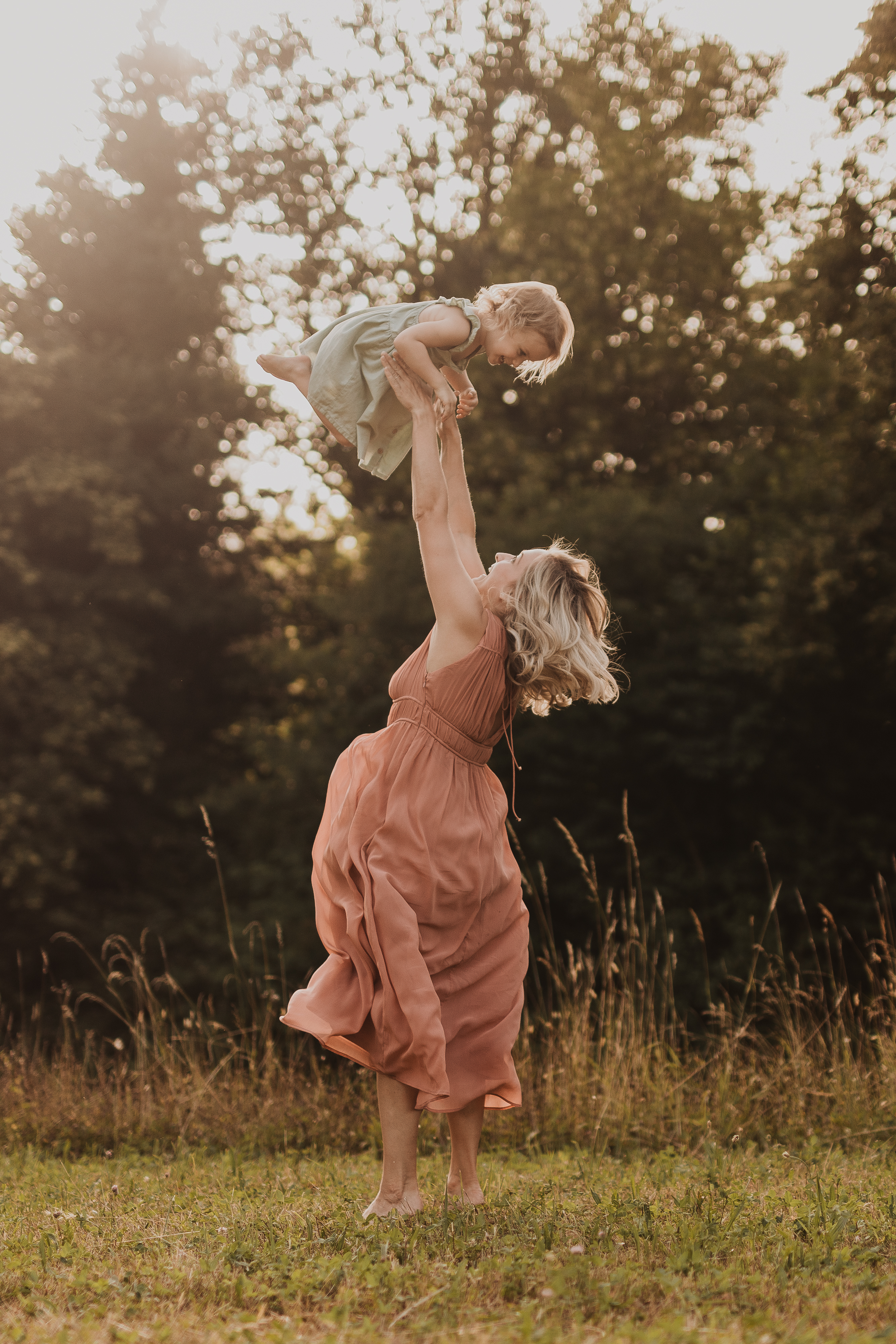 Eine Frau in einem fließenden pfirsichfarbenen Kleid hebt ein lächelndes kleines Kind über ihren Kopf. Sie stehen auf einer sonnenbeschienenen Wiese mit hohen Bäumen im Hintergrund. Diese von Natürliche Familienfotos aufgenommene Szene zeigt einen freudigen und lebhaften Moment zwischen ihnen und verkörpert die Essenz natürlicher Familienfotografie.