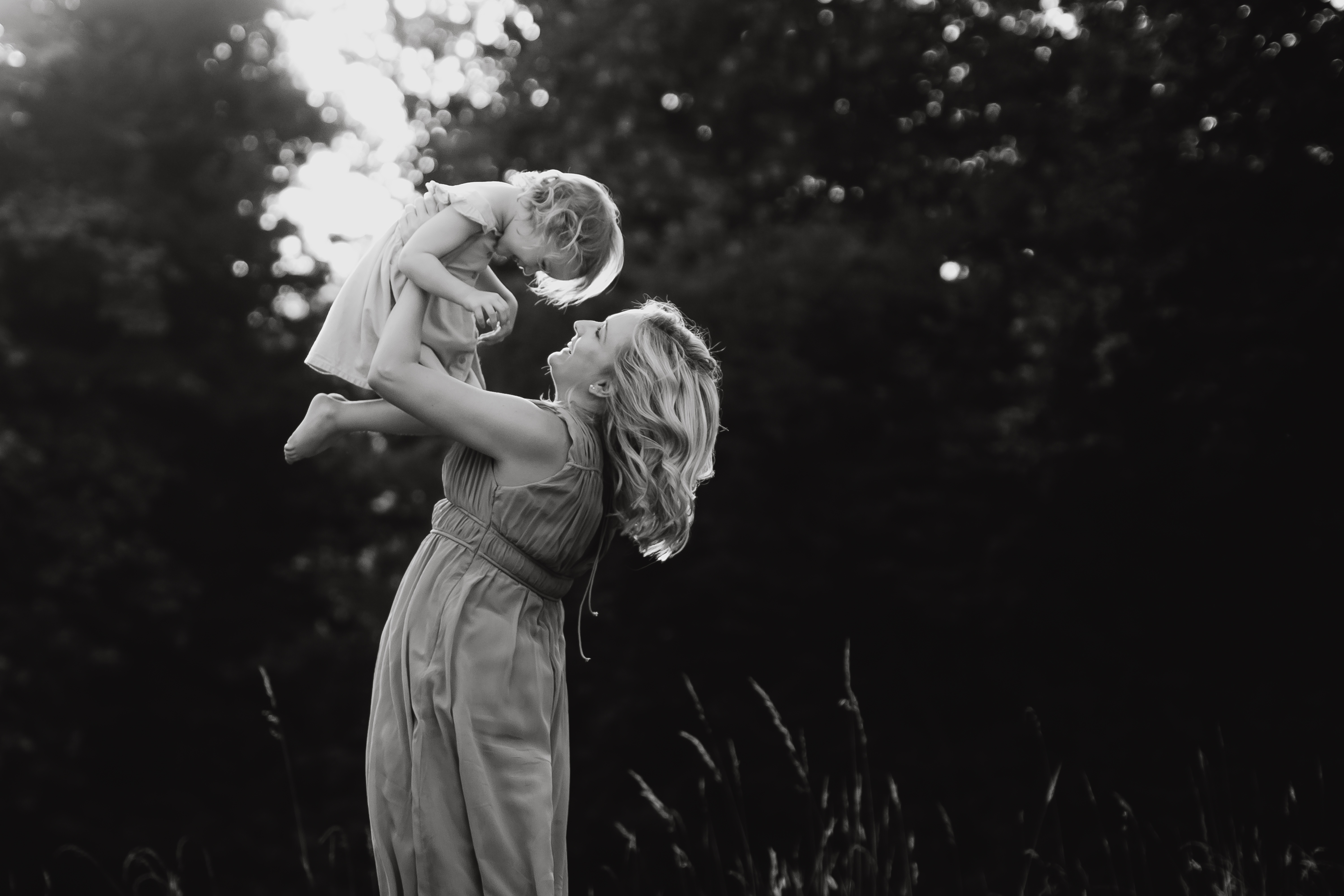 Eine Frau in einem ärmellosen Kleid hebt ein kleines Kind über ihren Kopf. Beide wirken fröhlich. Sie sind draußen auf einer Wiese mit Bäumen im Hintergrund. Dieses Familienshooting-Bild wurde von der Fotografin Tasha Ender aufgenommen und ist in Schwarzweiß gehalten. Das Sonnenlicht fällt durch die Bäume und verleiht ihnen einen warmen Schein.
