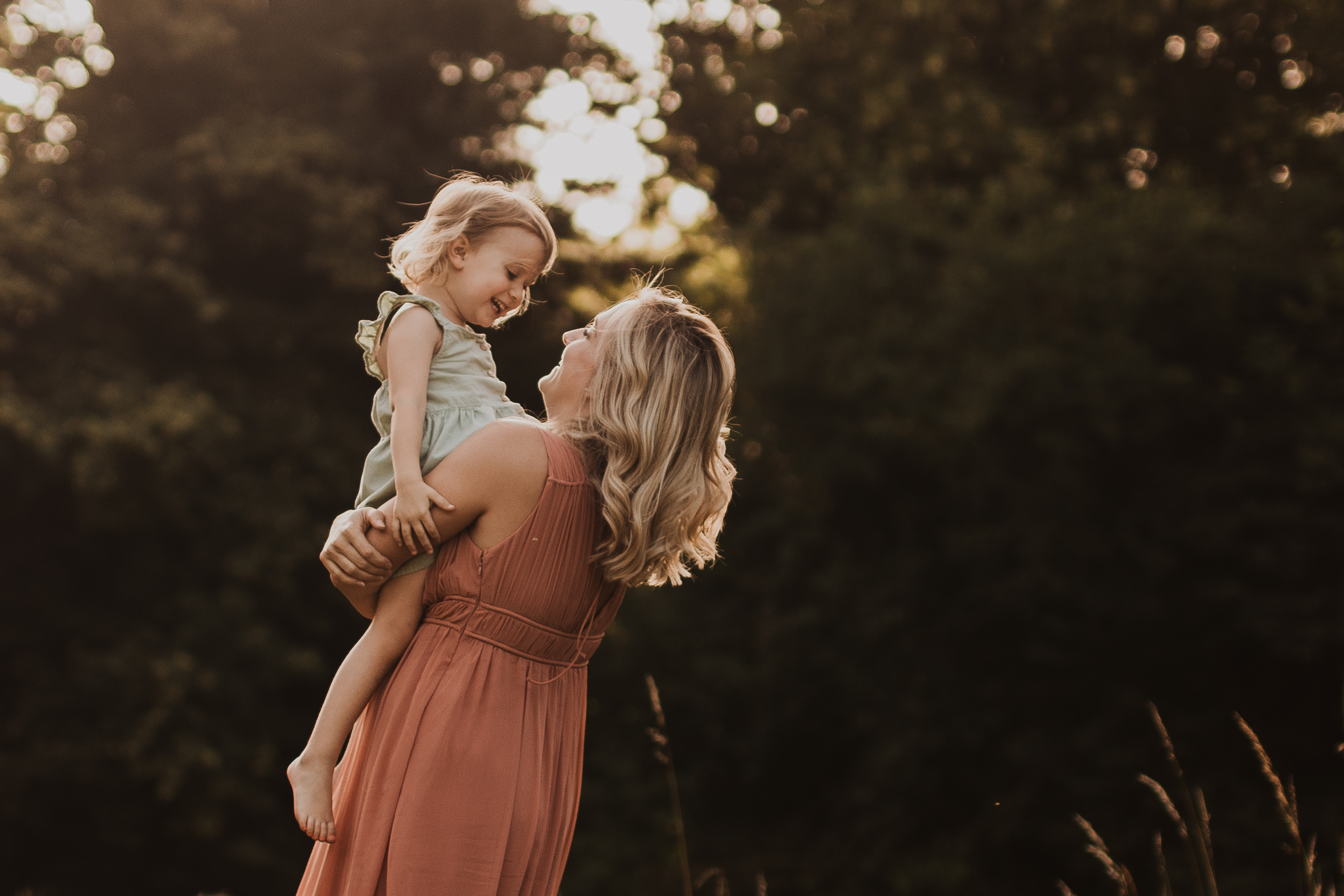 Eine Frau in einem pfirsichfarbenen Kleid, möglicherweise Teil eines Fotoshootings, hebt ein kleines Kind in einem hellgrünen Kleid hoch und hält es. Sie sind draußen in der Nähe von Baden, umgeben von hohem Gras und verschwommenem grünem Laub. Beide lächeln und blicken sich im warmen, natürlichen Licht an.