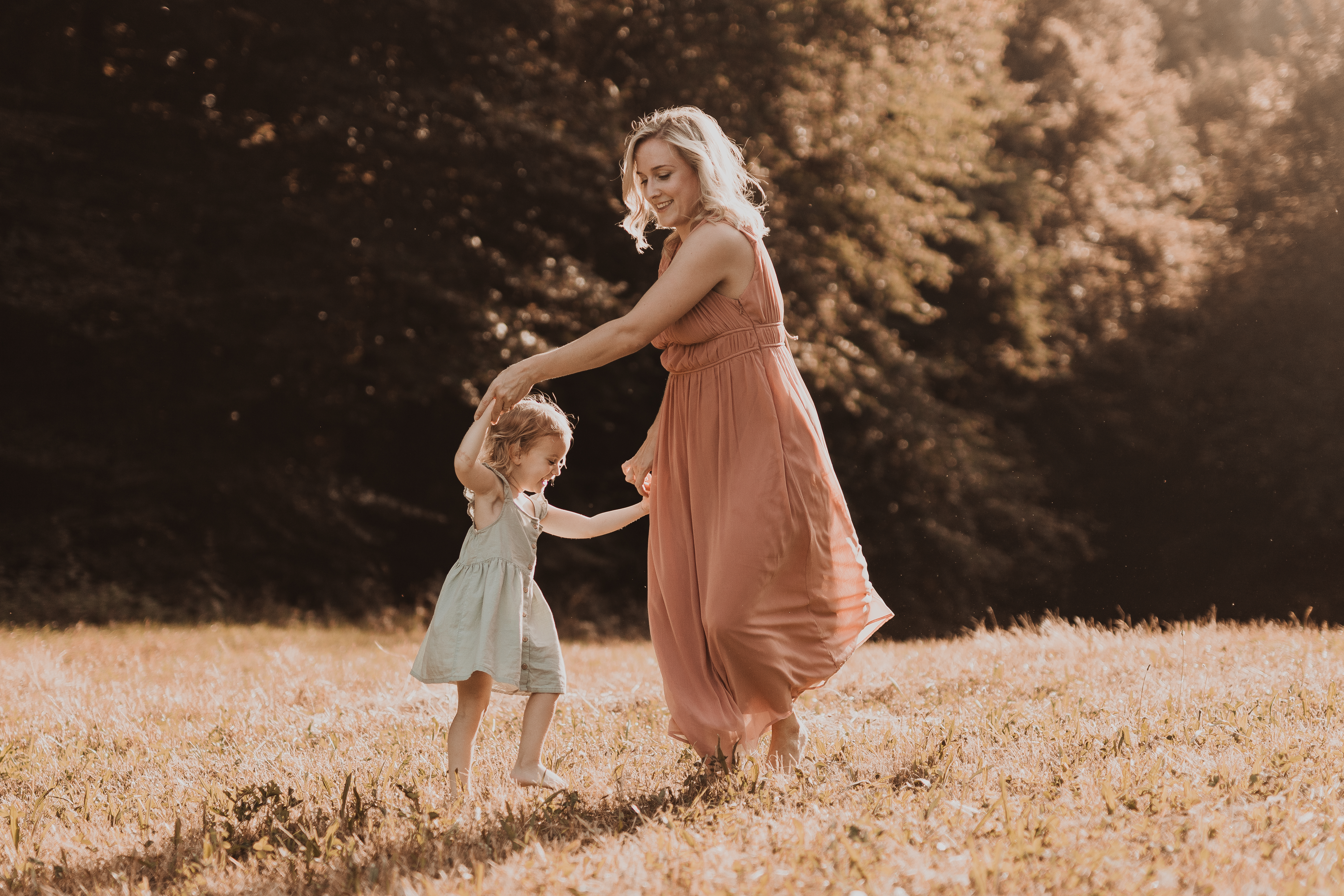 Eine junge Mutter im pastel rosa Kleid tanzt in der Abendsonne mit ihrer kleinen Tochter, die ein mintgrünes Kleidchen trägt. Die Fotografin hat den Moment der Freude zwischen Mutter und Tochter perfekt eingefangen.