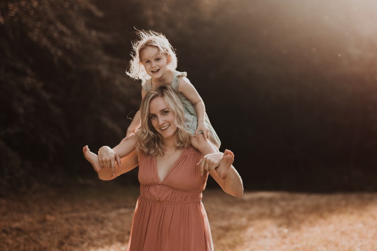 Eine Frau, wahrscheinlich Mutter, trägt ihr strahlendes kind auf den Schultern. Die Fotografin hat während dieses Mutter-Tochter Fotoshootings die warme Stimmung der golden hour genutzt um ein beosnders stimmungsvolles Bild zu kreieren.