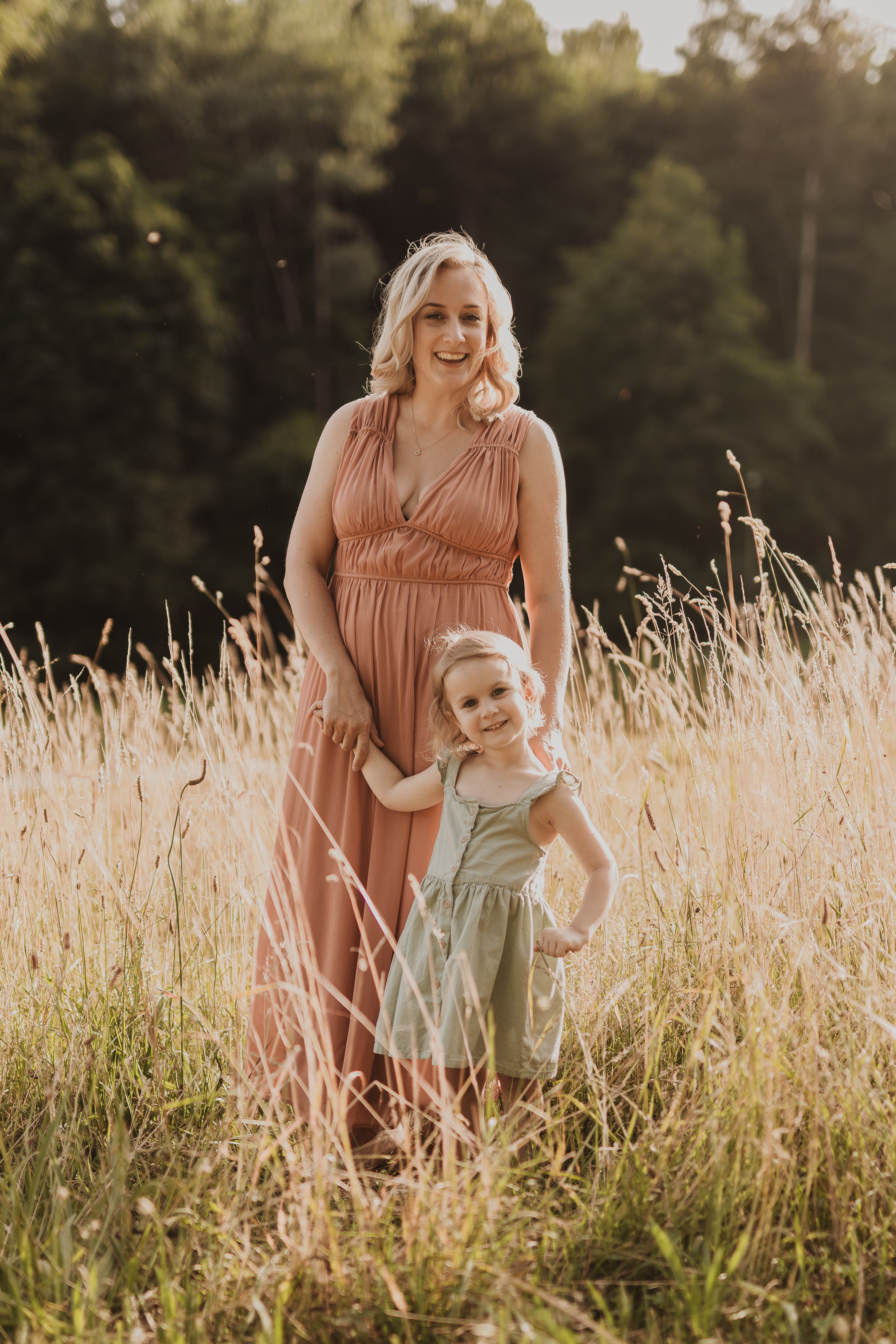Eine Frau in einem fließenden pfirsichfarbenen Kleid steht auf einer sonnigen Wiese und hält ein junges Mädchen in einem hellgrünen Kleid an der Hand. Sie lächeln sich herzlich an und verkörpern die Freude, die unsere auf natürliche Familienfotos spezialisierte Fotografin eingefangen hat. Bäume und sanftes Sonnenlicht bilden dabei die perfekte Kulisse.