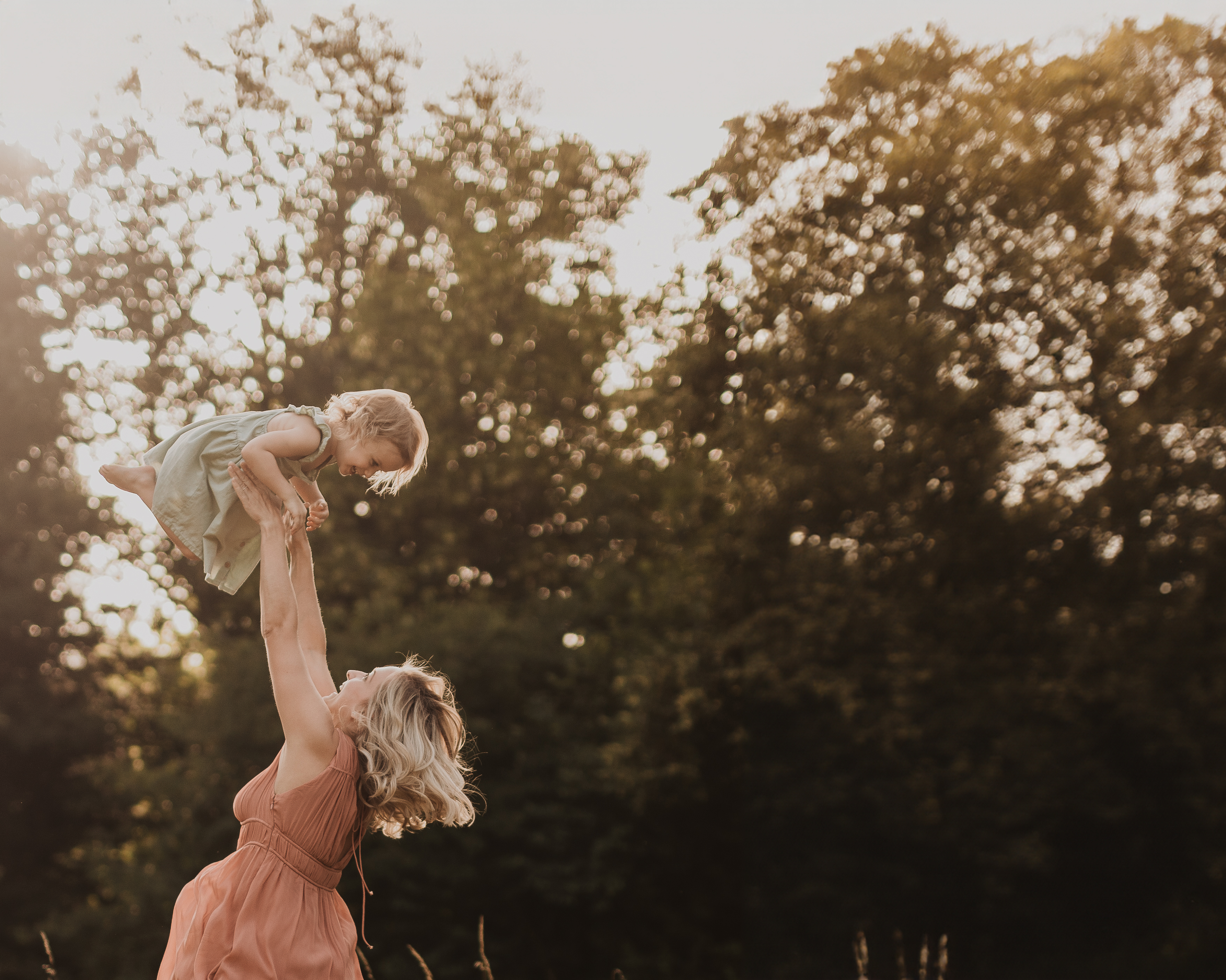 Bei einem Fotoshooting hebt eine Frau in einem pfirsichfarbenen Kleid ein kleines Kind in einem grünen Kleid hoch in die Luft. Sie befinden sich draußen in einem Park oder Waldgebiet mit Bäumen im Hintergrund und Sonnenlicht, das durch die Blätter fällt und eine warme, goldene Atmosphäre schafft. Beide scheinen glücklich zu sein.