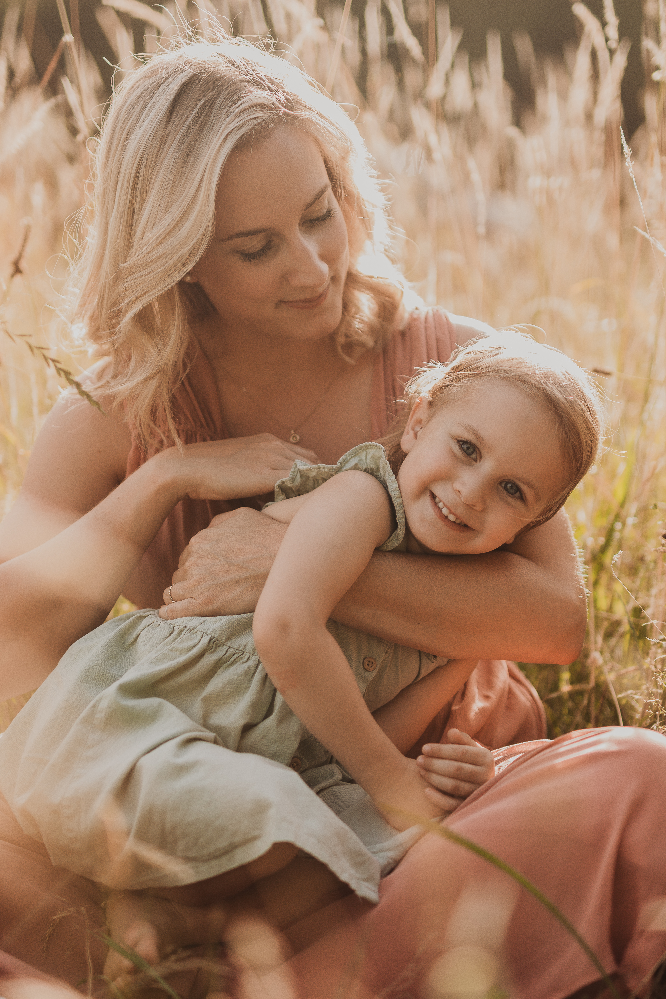Eine Frau mit blondem Haar sitzt in einem sonnenbeschienenen Feld mit hohem Gras und hält ein kleines Kind auf ihrem Schoß. Das Kind in einem hellen Kleid lächelt herzlich in die Kamera, während die in Pfirsich gekleidete Frau sie liebevoll ansieht – ein zarter Moment, eingefangen von der Fotografin Tasha Ender.