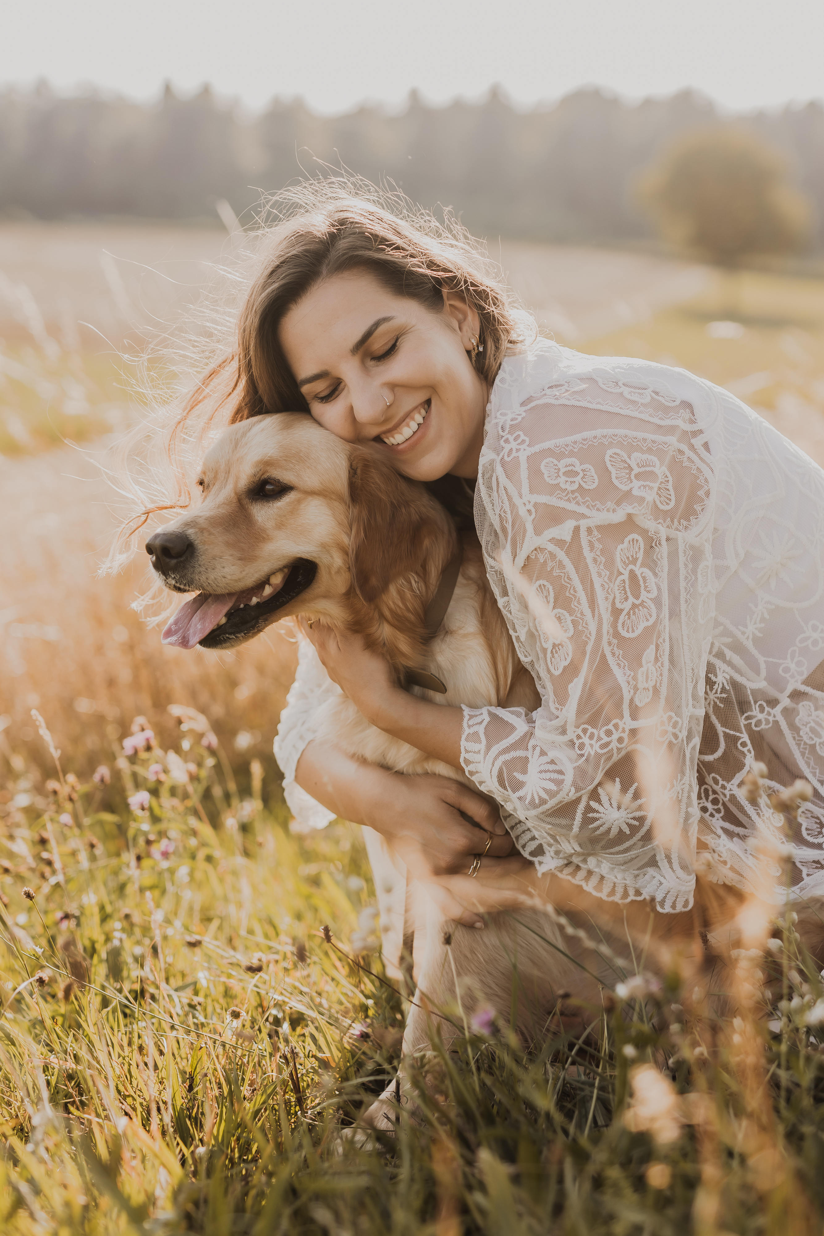 Eine Frau in einem weißen Spitzenkleid hockt auf einer Wiese und umarmt ihren Golden Retriever. Beide lächeln und wirken glücklich. Die Boho-inspirierte Szene ist voller Wildblumen und in warmes Sonnenlicht getaucht. Im Hintergrund sind Bäume zu sehen – perfekt für jeden Tierfotografen.