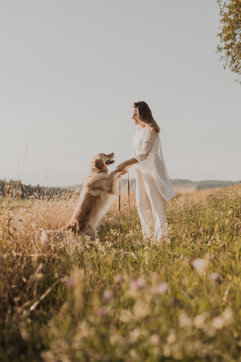Eine Frau in einem weißen Boho-Kleid steht in einem Feld mit hohem Gras und Wildblumen und hält die Vorderpfoten eines stehenden Golden Retrievers. Der Hund scheint mitten im Spiel zu sein und die Frau lächelt. Der Himmel ist klar und die Szene ist in warmes Licht getaucht, als ob sie von einem erfahrenen Hundefotografen aufgenommen worden wäre.