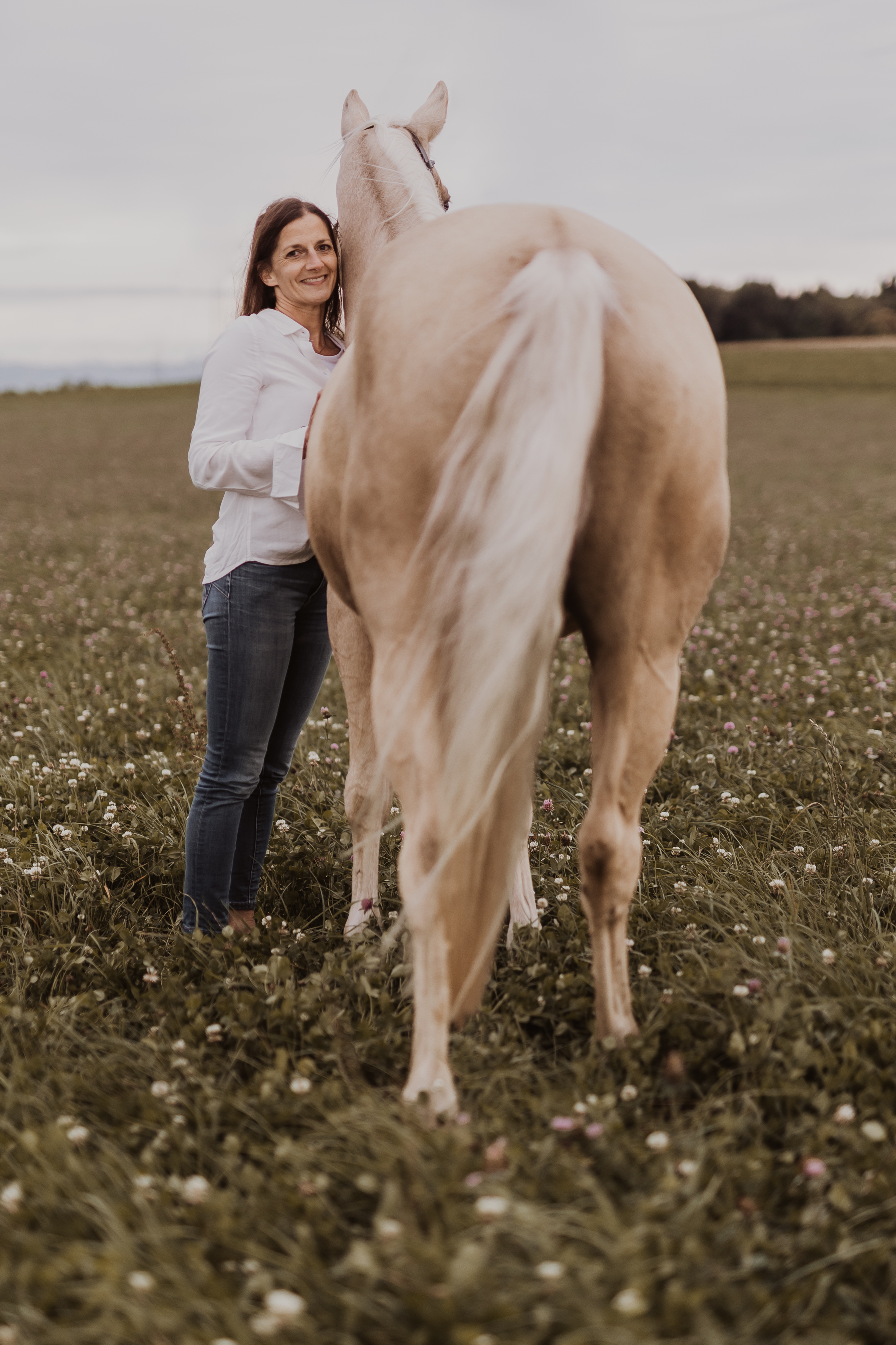 Eine Person mit langen Haaren, die ein weißes Hemd und Jeans trägt, steht neben einem hellen Pferd auf einer weiten, grünen, mit Blumen übersäten Wiese. Die Szene, die vom Fotoshooting-Service von Tasha Ender Photography aufgenommen wurde, zeigt die Person gegenüber dem Pferd, das seinen Schweif abgewandt hat. Der bewölkte Himmel verleiht dem ruhigen Moment sanftes Licht.