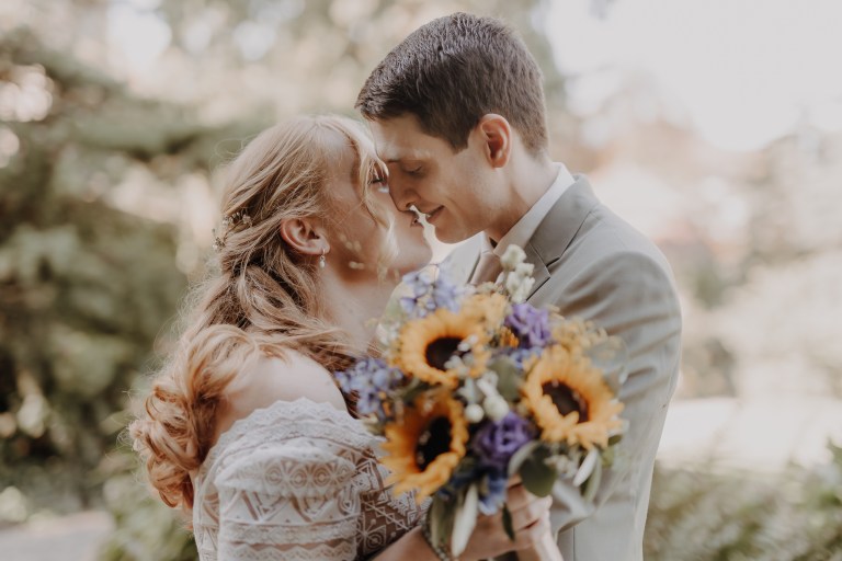 Ein Paar steht eng beieinander, berührt seine Stirn und lächelt, und teilt einen intimen Moment während der Hochzeitsfotografie von Tasha Ender Photography. Die Frau hält einen farbenfrohen Strauß aus Sonnenblumen und lila Blumen, während der Mann einen hellen Anzug trägt. Im Hintergrund ist der üppige, sonnenbeschienene Garten der Villa Boveri in Baden.