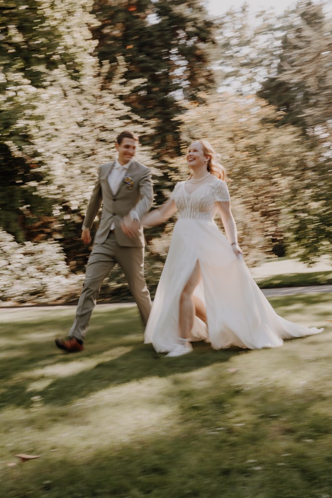 Ein Paar in Hochzeitskleidung hält fröhlich Händchen, während es durch einen üppigen, sonnenbeschienenen Park spaziert. Die Braut trägt ein fließendes weißes Kleid und der Bräutigam einen hellgrauen Anzug. Beide wirken glücklich, lächeln und schauen sich während ihres Hochzeitsreportage-Fotoshootings von Tasha Ender inmitten des Grüns an.