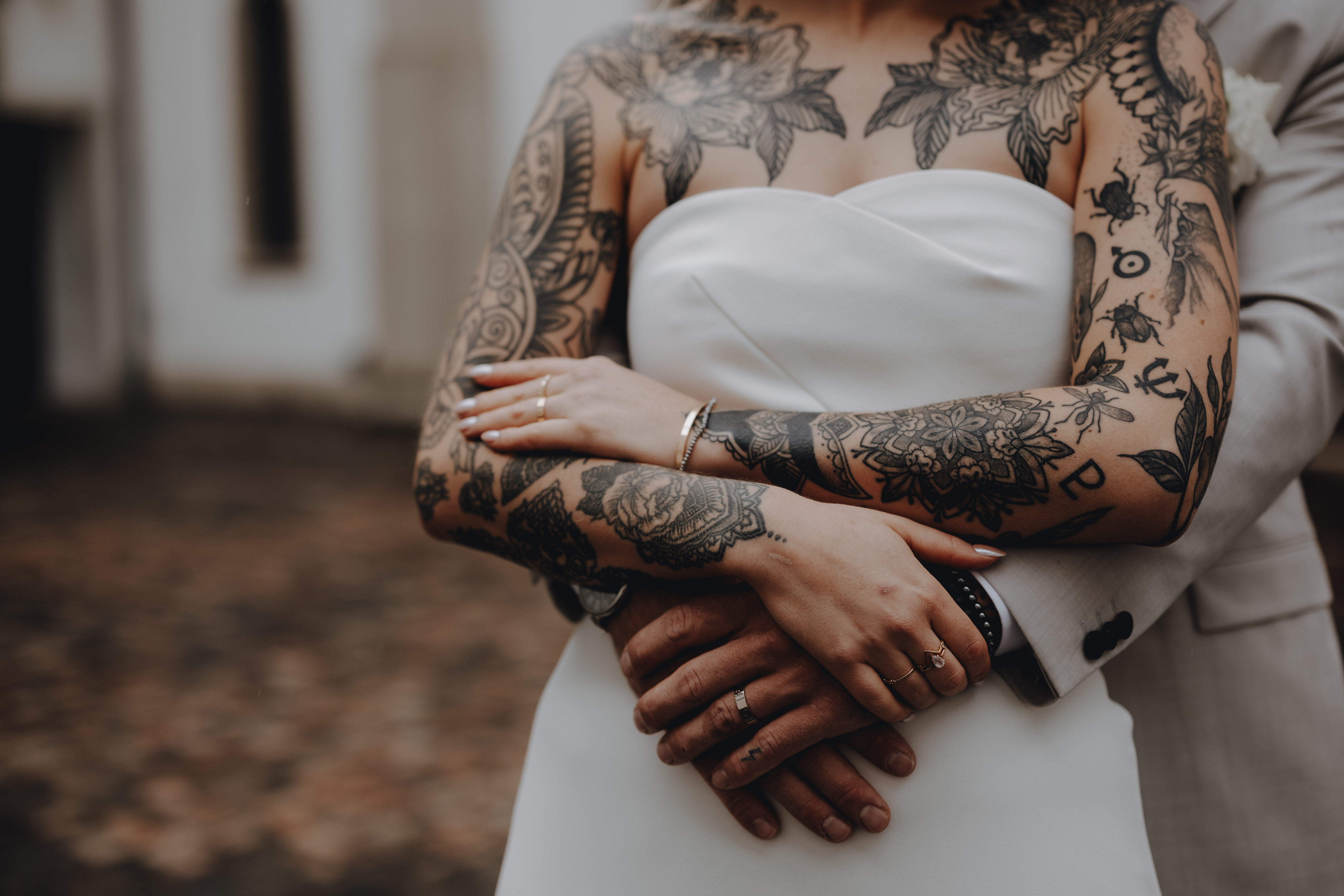 Eine Nahaufnahme eines Paares, das sich während eines Fotoshootings umarmt. Die Person vorne, die ein trägerloses weißes Kleid trägt, zeigt umfangreiche und aufwendige Tattoos auf Armen und Oberkörper. Die Person dahinter, die einen hellen Anzug trägt, hat ihre Arme um die andere geschlungen. Wunderschön aufgenommen von Hochzeitsfotografin Tasha Ender.