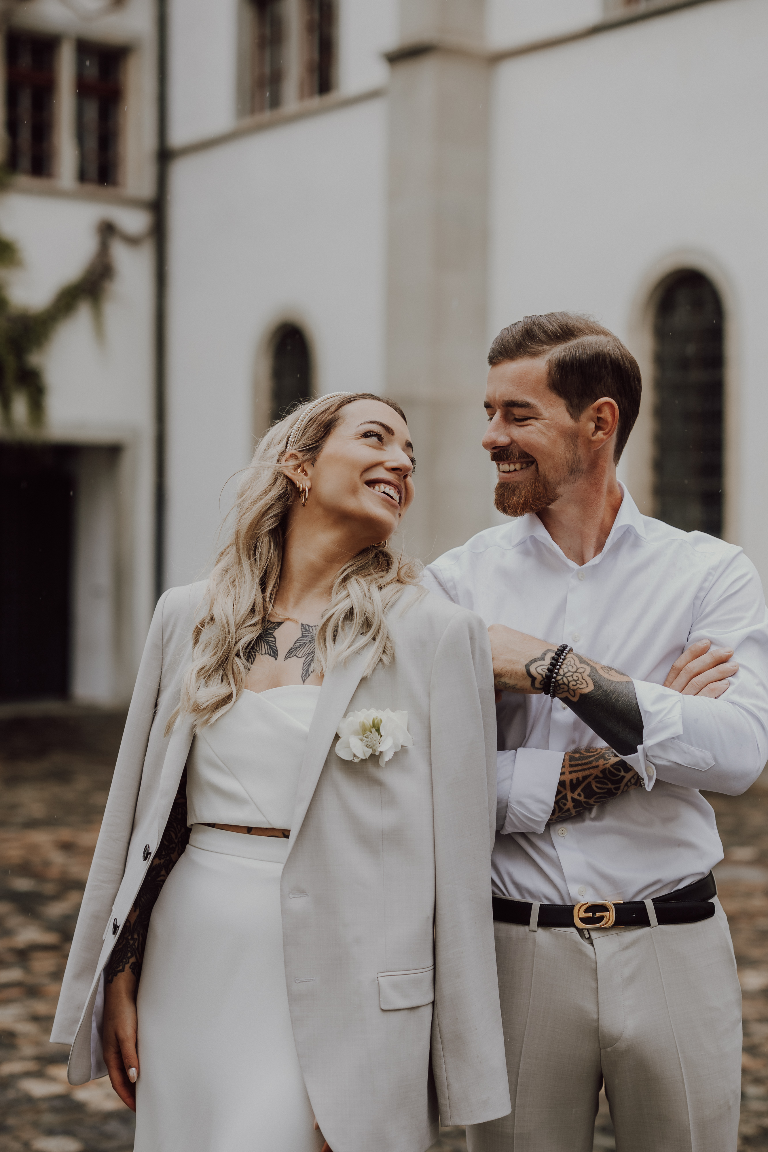 Ein lächelndes Hochzeitspaar steht dicht beieinander vor einem Gebäude mit Bogenfenstern. Die Frau mit blonden Haaren und Tattoos trägt ein weißes Kleid mit einem beigefarbenen Blazer über den Schultern. Der Mann mit einem ordentlich gestutzten Bart trägt ein weißes Hemd und beigefarbene Hosen.