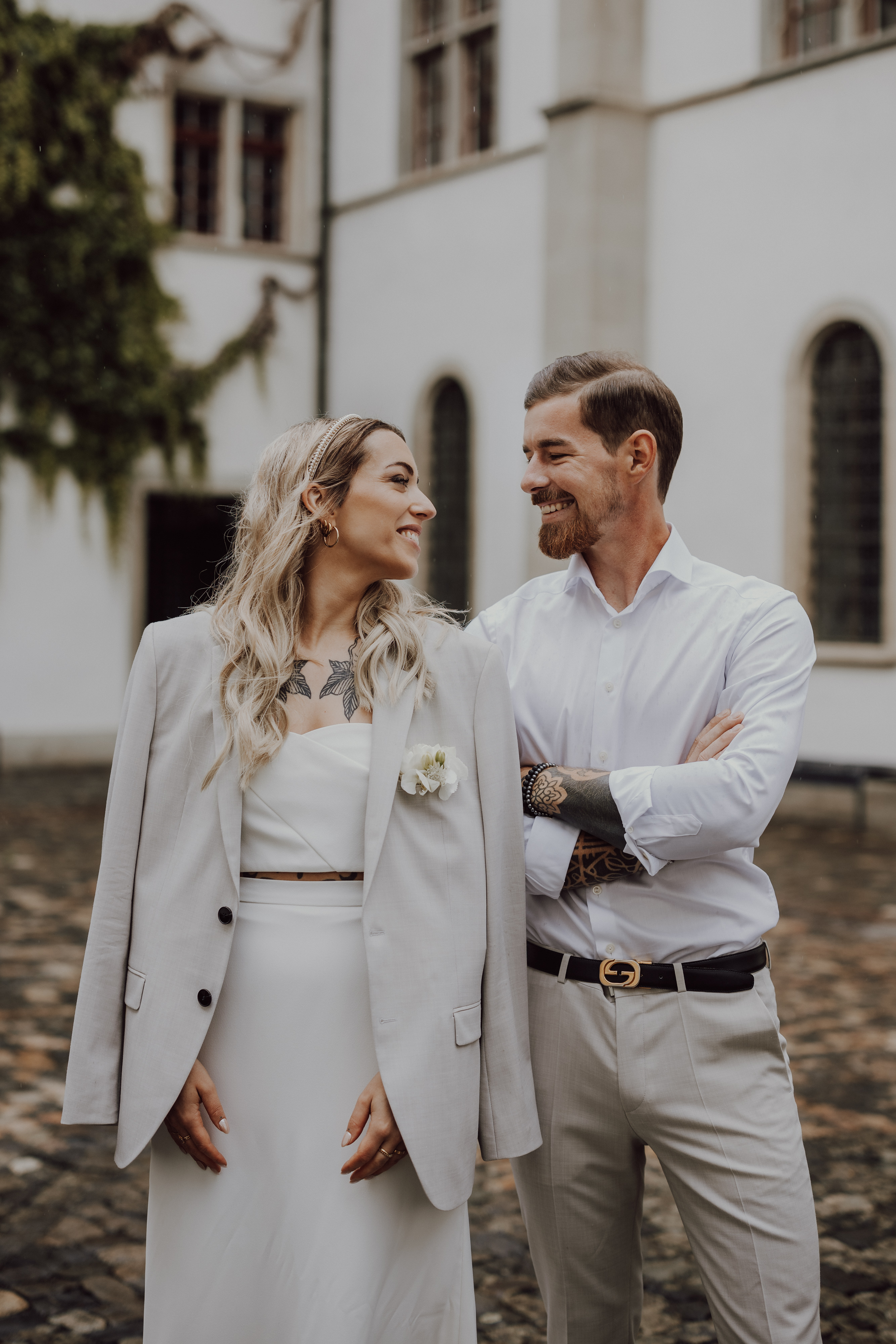 Ein Hochzeitspaar steht vor einem Gebäude mit weißen Wänden und lächelt sich an. Die Frau hat lange blonde Haare und trägt einen hellgrauen Blazer über einem weißen Kleid. Der Mann hat kurze Haare, einen Bart und trägt ein weißes Oberhemd und hellgraue Hosen, mit verschränkten Armen.