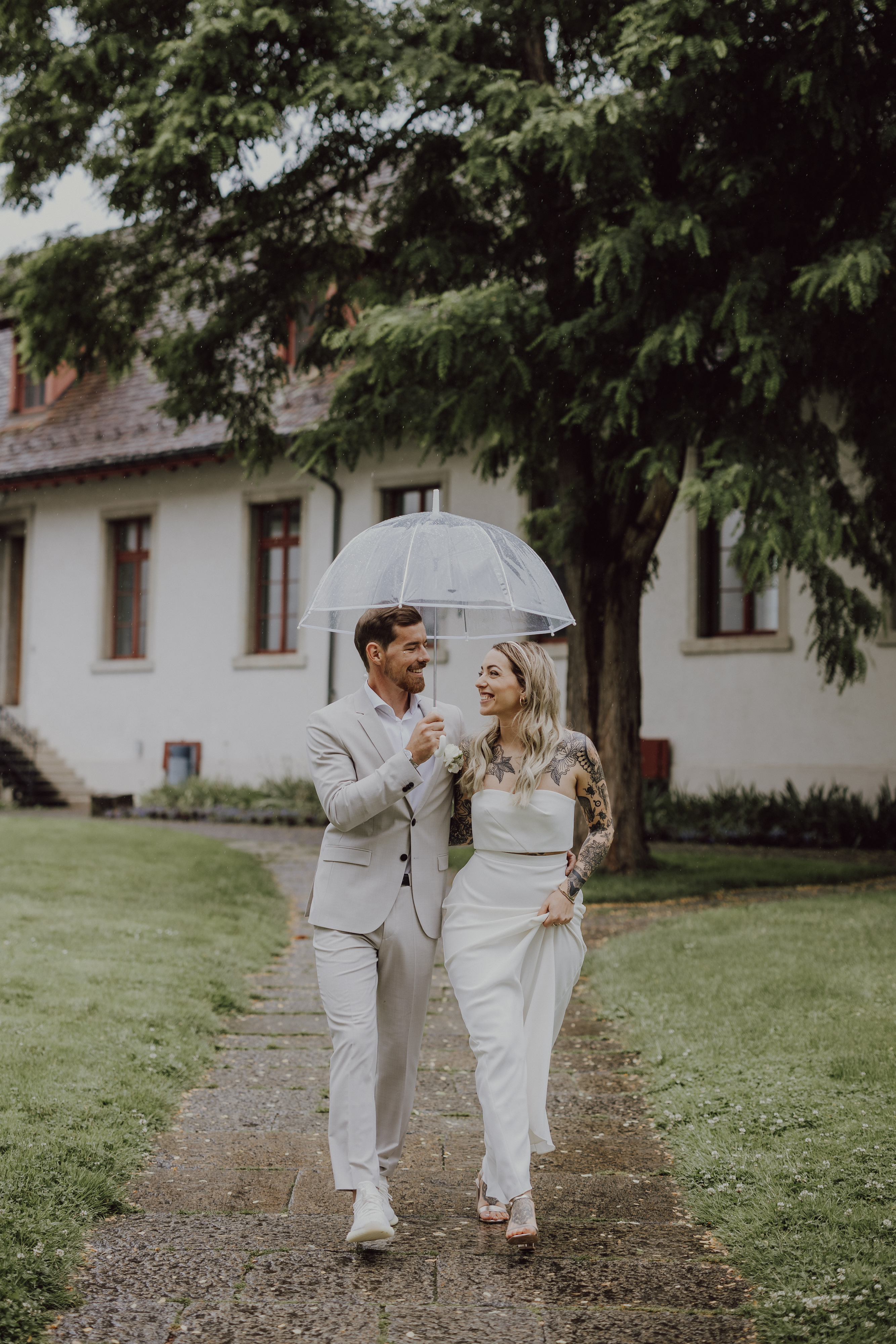 Ein Hochzeitspaar geht an einem regnerischen Tag Arm in Arm spazieren, teilt sich einen transparenten Regenschirm und lächelt sich an. In formeller Kleidung, der Mann in einem hellgrauen Anzug und die Frau in einem weißen Kleid, schlendern sie an Gebäuden und Bäumen vorbei. Perfekt für jede Hochzeitsfotografin!