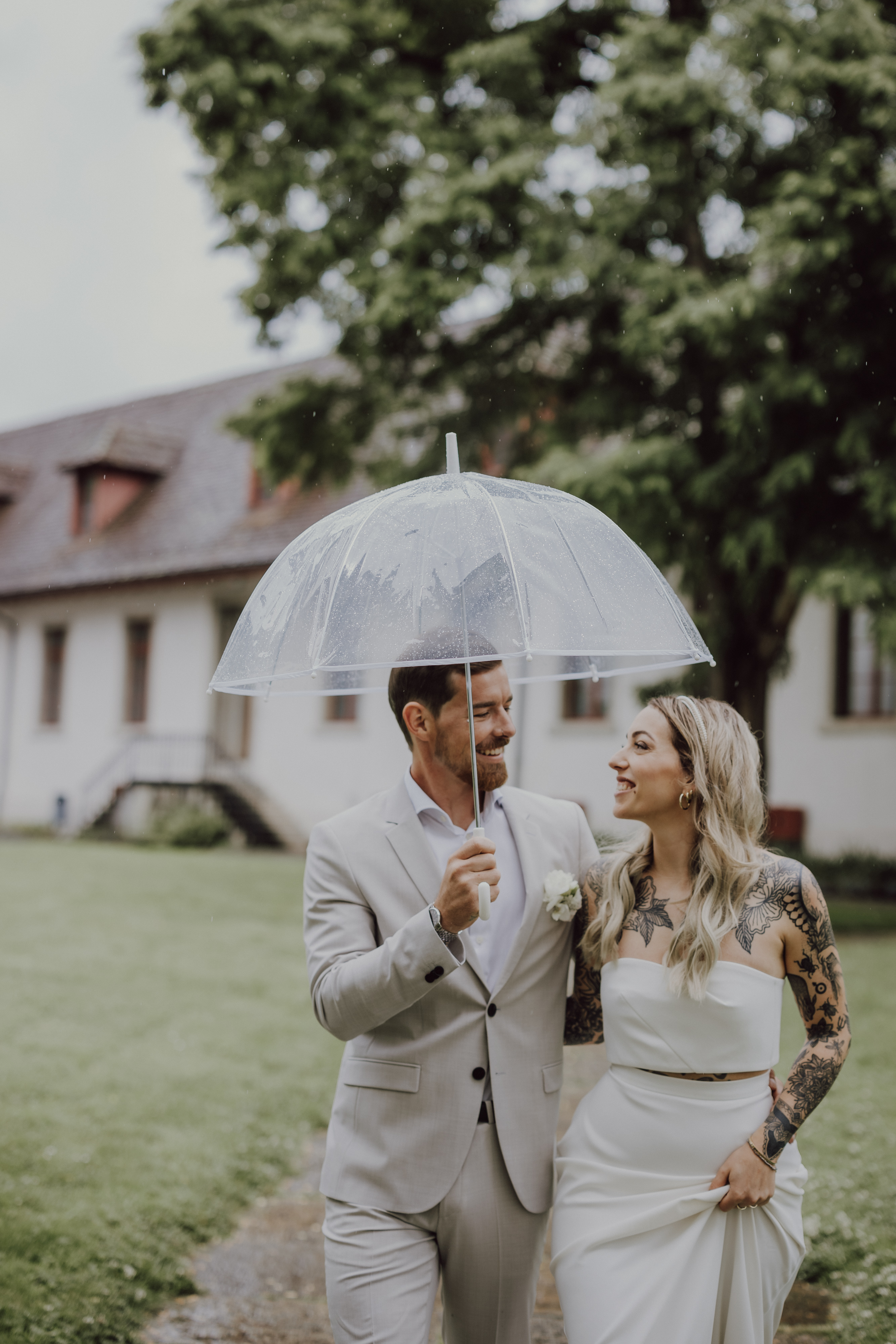 Ein frisch vermähltes Paar (Hochzeitspaar) geht an einem regnerischen Tag gemeinsam im Freien spazieren. Der Bräutigam in einem hellgrauen Anzug hält einen durchsichtigen Regenschirm, während die Braut in einem weißen Kleid mit Tätowierungen auf dem linken Arm ihn anlächelt. Sie stehen vor einem Gebäude mit Bäumen im Hintergrund.