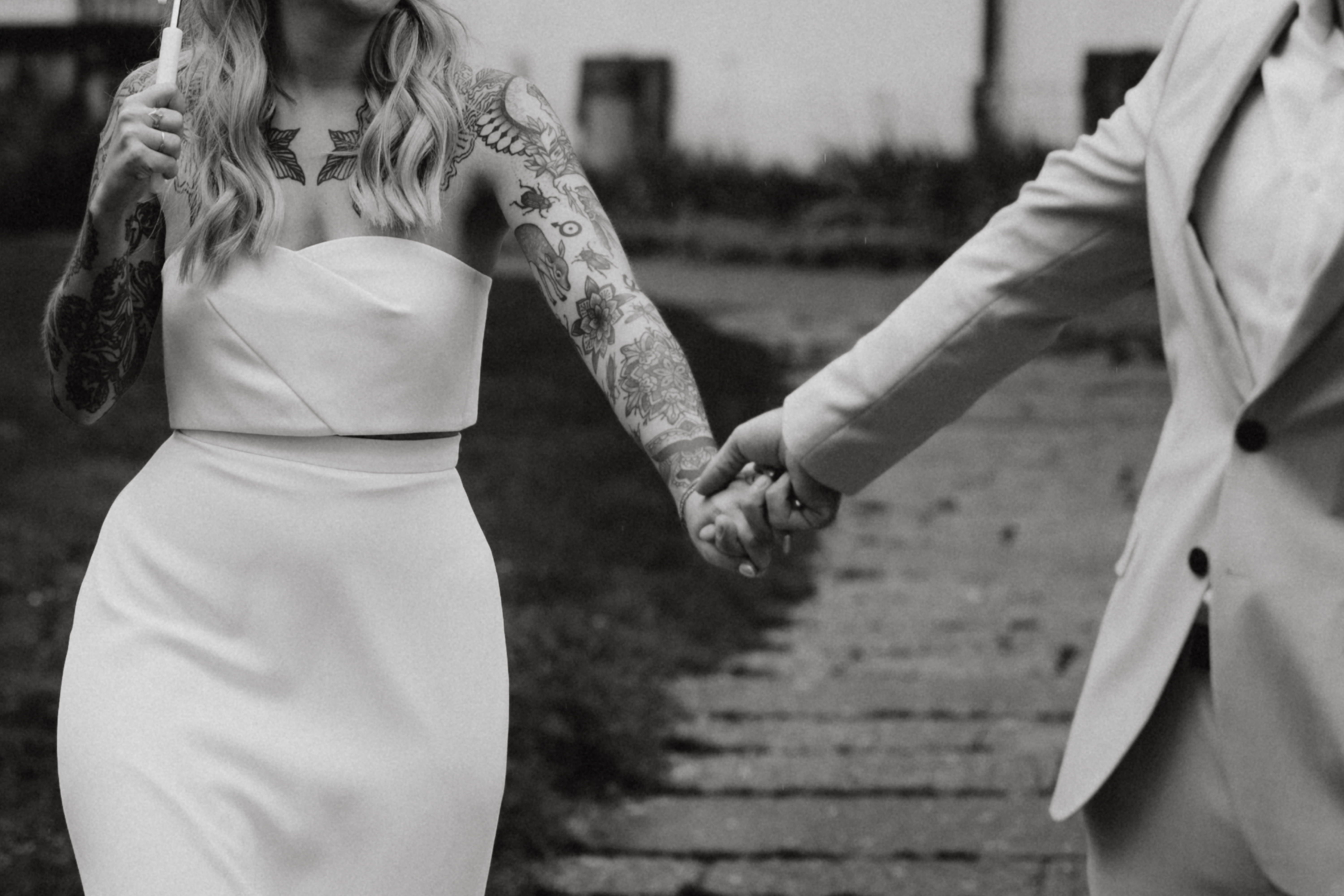 Ein Schwarzweißfoto zeigt eine tätowierte Person in einem schulterfreien Kleid, die Händchen hält mit einer anderen Person im Anzug, die auf einem Steinweg geht. Nur ihre Oberkörper und Arme sind sichtbar und fangen den Moment der Intimität und Verbundenheit des Hochzeitspaares ein.
