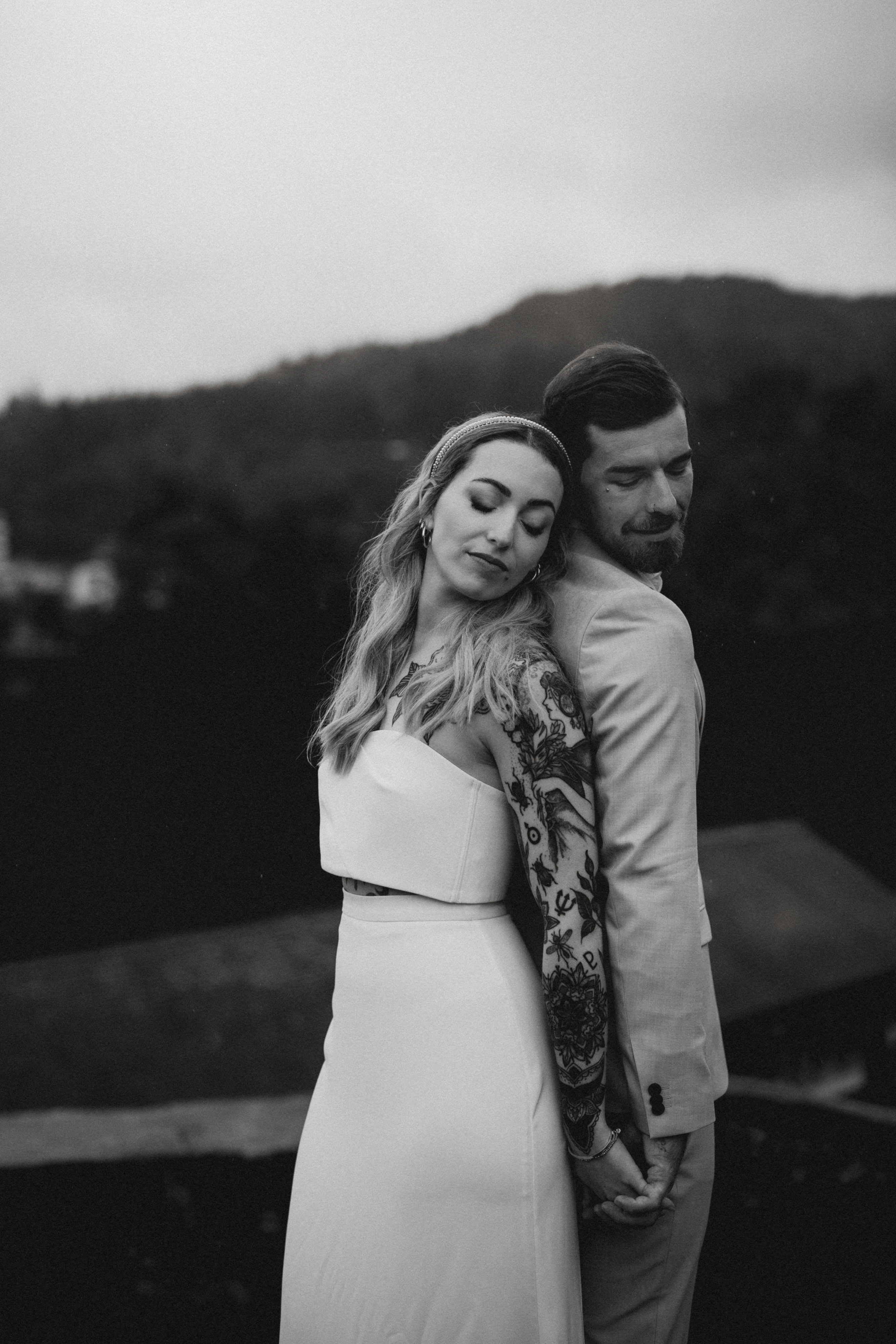 Ein Schwarzweißfoto aus der Fotoshooting-Sitzung von Tasha Ender Photography zeigt ein Paar, das an seinem Hochzeitstag Rücken an Rücken steht. Die Braut mit langem, welligem Haar und tätowiertem Arm ist elegant in ein ärmelloses Kleid gekleidet. Der Bräutigam mit kurzen Haaren und Bart ergänzt seinen Look mit einem hellen Anzug. Sie posieren auf einer Außenterrasse mit atemberaubenden Bergen im Hintergrund.