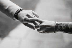 Nahaufnahme eines Schwarzweißfotos von Tasha Ender, auf dem zwei Menschen ihre Finger berühren. Eine Person hat einen tätowierten Arm, der mit Ringen und einem Armband geschmückt ist, während die andere ein helles Hemd und ein Perlenarmband trägt. Der Hintergrund ist unscharf, was diesem Meisterwerk der Hochzeitsfotografin Tiefe verleiht.