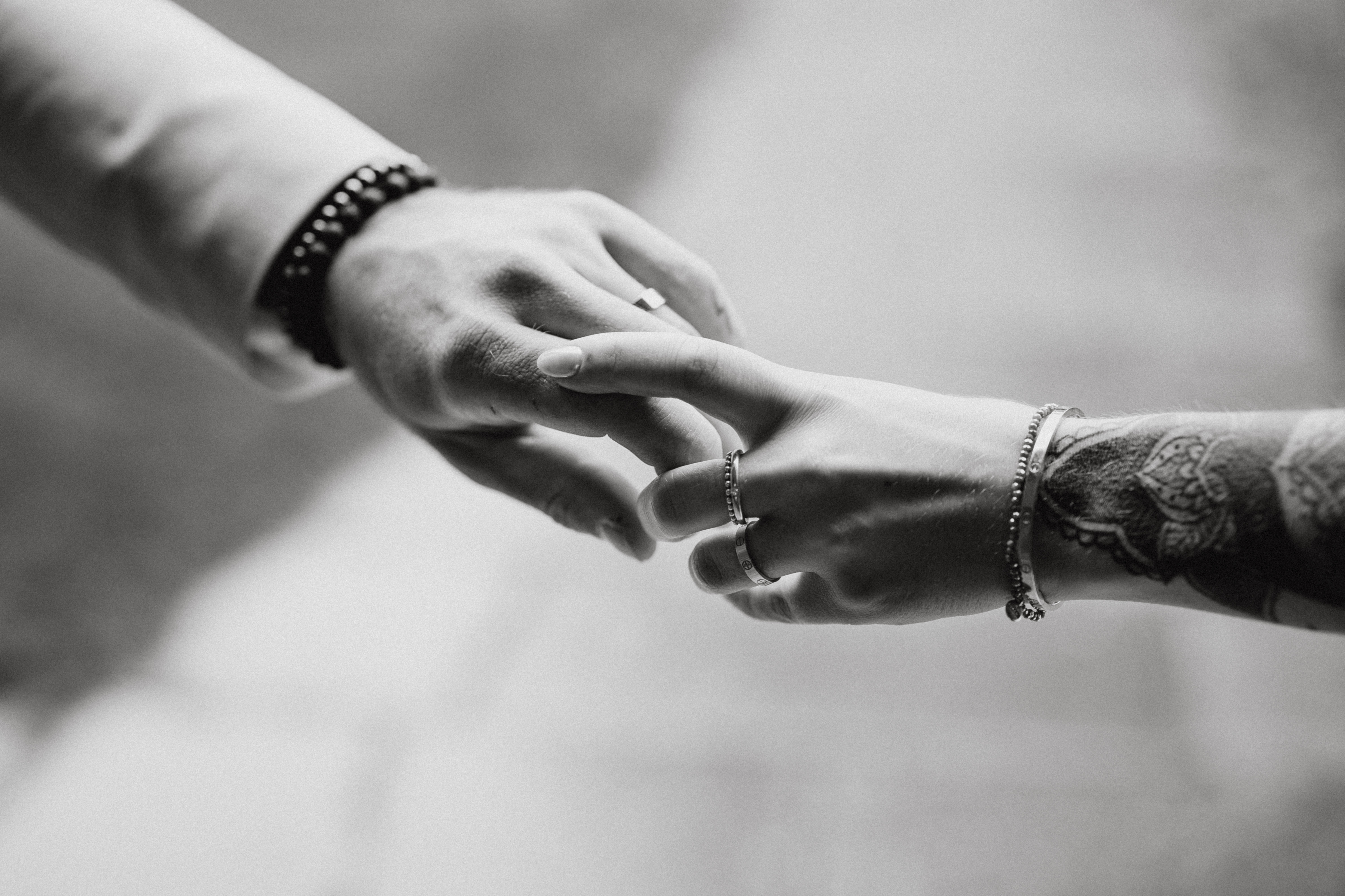 Nahaufnahme eines Schwarzweißfotos von Tasha Ender, auf dem zwei Menschen ihre Finger berühren. Eine Person hat einen tätowierten Arm, der mit Ringen und einem Armband geschmückt ist, während die andere ein helles Hemd und ein Perlenarmband trägt. Der Hintergrund ist unscharf, was diesem Meisterwerk der Hochzeitsfotografin Tiefe verleiht.