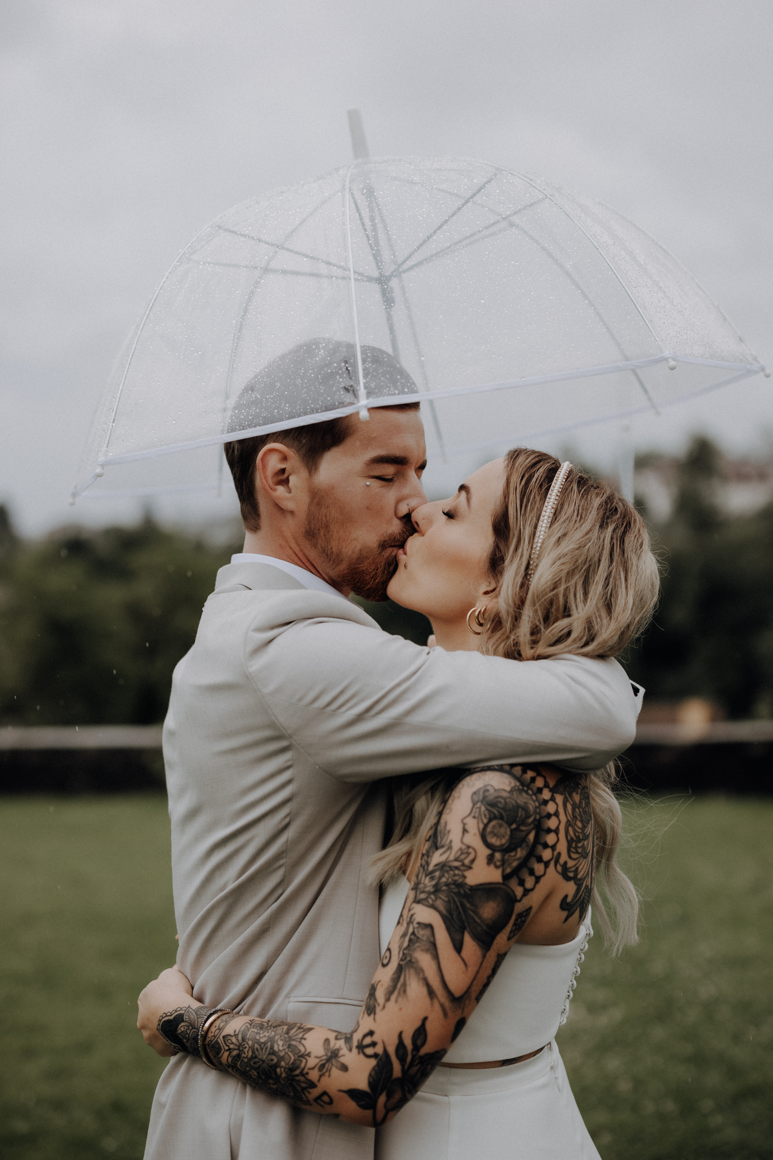 Ein Paar in Hochzeitskleidung küsst sich an einem regnerischen Tag unter einem durchsichtigen Regenschirm. Die Braut mit tätowierten Armen und geschlossenen Augen wird eng von dem Bräutigam im hellen Anzug gehalten. Wunderschön von Hochzeitsfotografin Tasha Ender fotografiert, stehen sie draußen mit einer verschwommenen grünen Landschaft im Hintergrund.