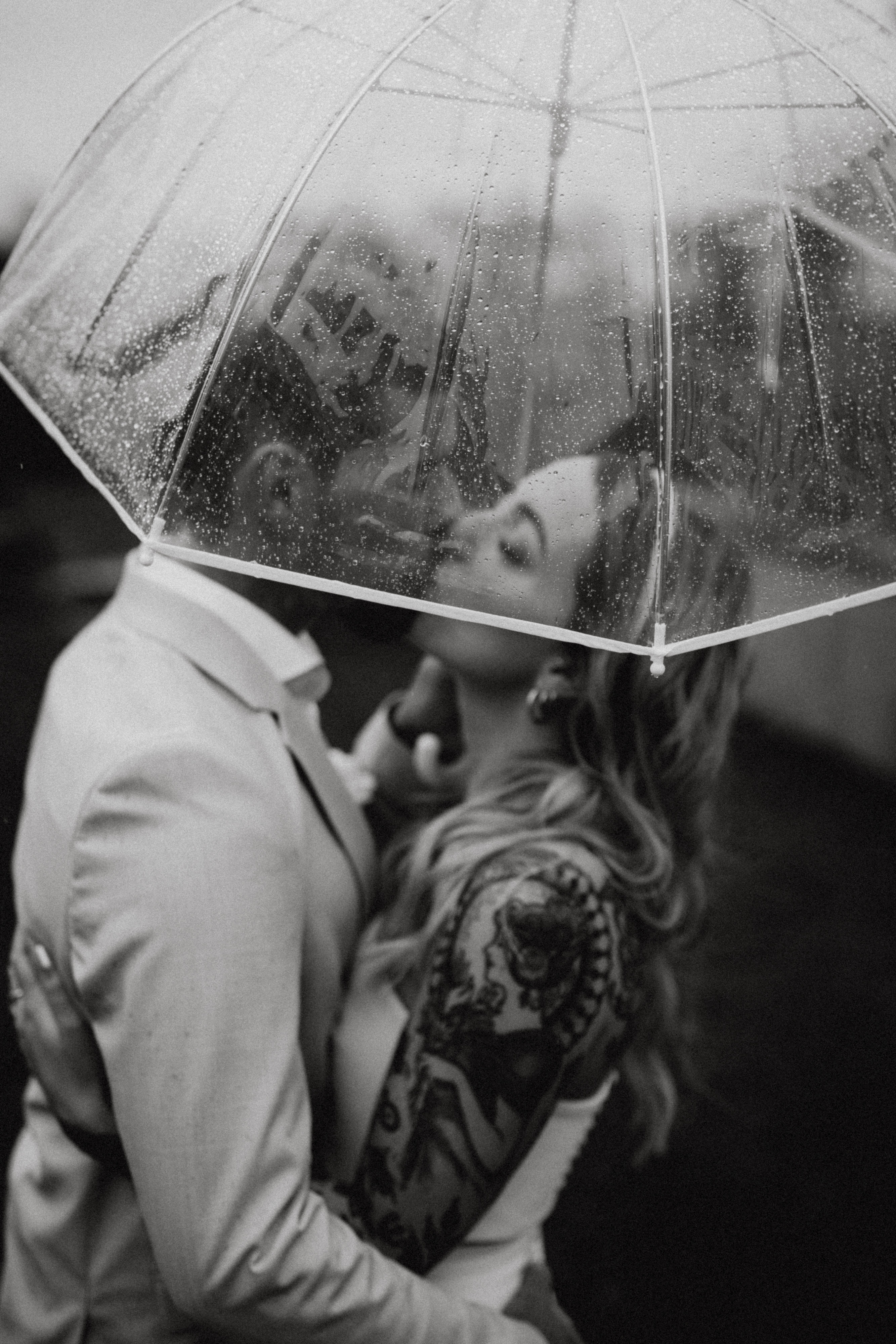Ein Schwarz-Weiß-Foto von Hochzeitsfotografin Tasha Ender zeigt ein Hochzeitspaar, das sich an einem regnerischen Tag unter einem durchsichtigen Regenschirm küsst. Regentropfen zieren den Schirm, während sich der Mann im Anzug und die Frau mit auffälligen Armtattoos im ärmellosen Kleid eng umarmen.