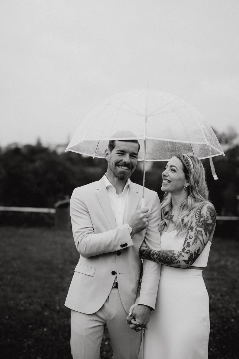 Ein Paar in Hochzeitskleidung steht im Freien, hält Händchen und lächelt unter einem durchsichtigen Regenschirm. Der Mann trägt einen hellen Anzug, während die Frau ein weißes Kleid trägt, das ihre sichtbaren Tattoos zur Schau stellt. Der Hintergrund ist eine verschwommene Naturlandschaft, perfekt für atemberaubende Hochzeitsfotos.
