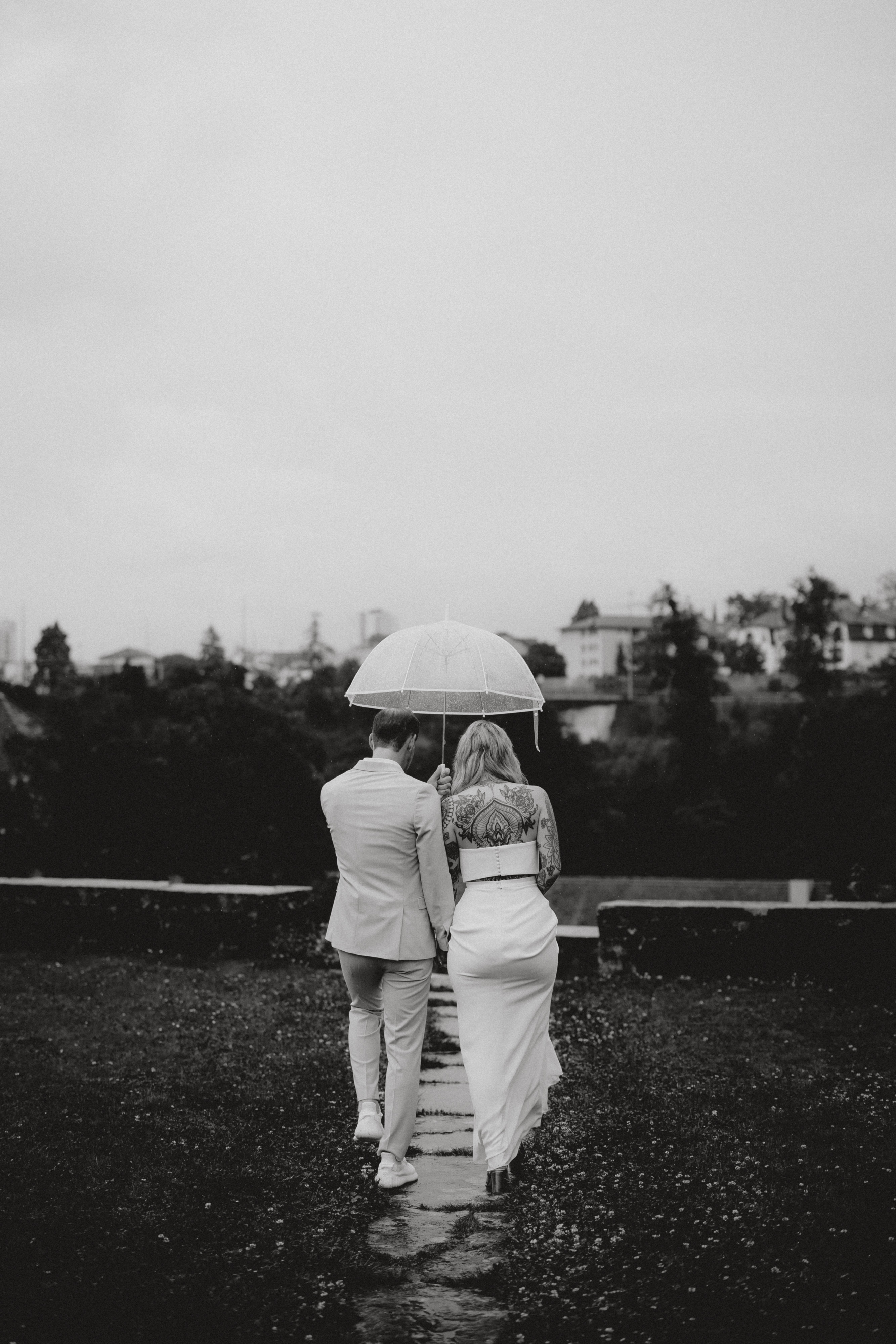 Ein Schwarz-Weiß-Foto aus der Hochzeitsfotografie-Kollektion von Tasha Ender Photography zeigt ein Paar, das auf einem Kiesweg in Baden spazieren geht, eng aneinander gekuschelt unter einem gemeinsamen Sonnenschirm. Der Mann, gekleidet in einen hellen Anzug, und die Frau im schulterfreien Kleid gehen gemeinsam vor einer malerischen Kulisse aus Bäumen und Gebäuden – ein eleganter Moment während ihres Fotoshootings im Aargau.