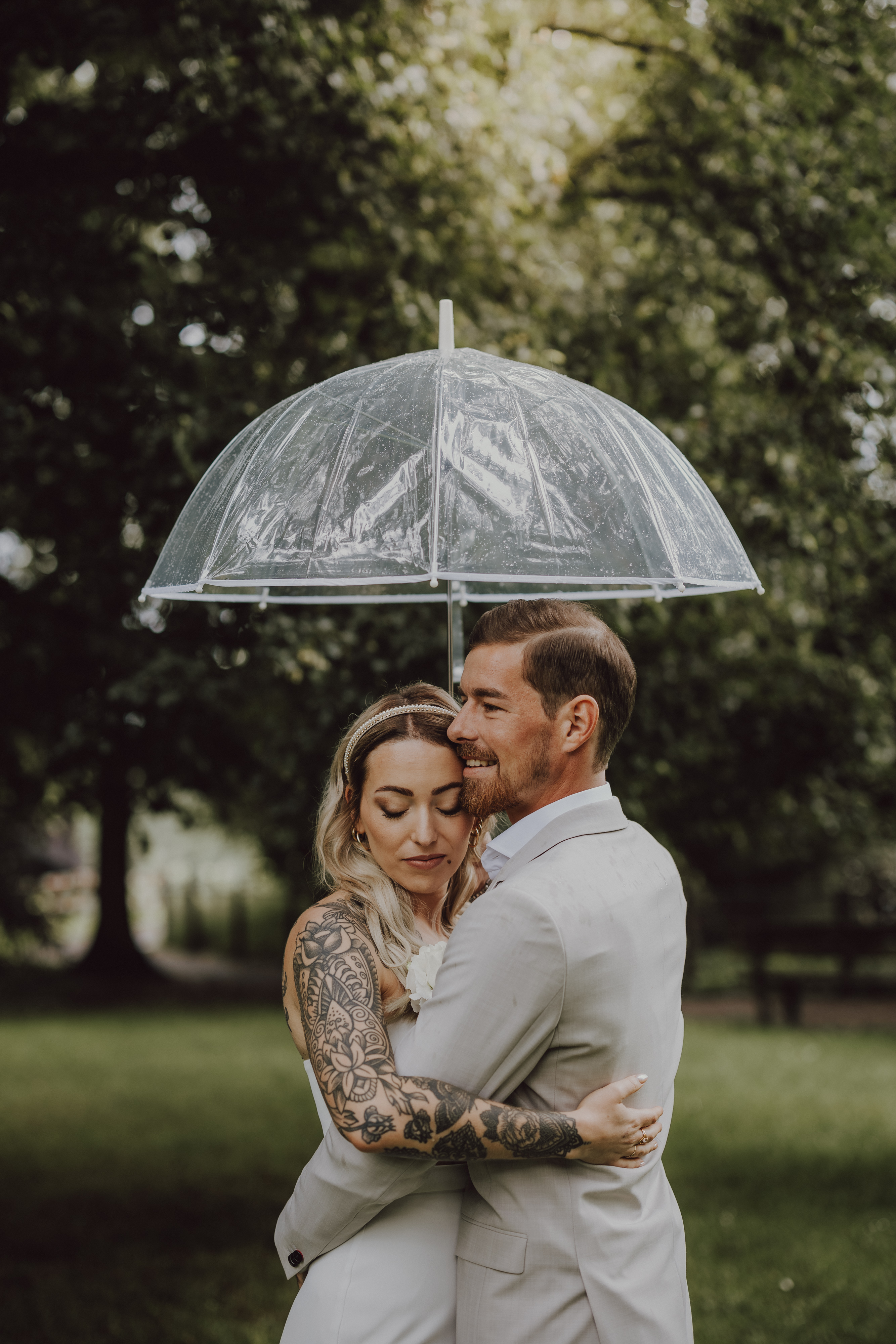 Ein für die Hochzeit gekleidetes Paar umarmt sich unter einem transparenten Regenschirm. Die Braut, deren Tattoos sichtbar sind, hat die Augen geschlossen, während der Bräutigam lächelt und sie festhält. Sie stehen auf einer Wiese mit üppigen grünen Bäumen im Hintergrund und fangen trotz des leichten Regens den perfekten Moment während ihrer Hochzeitsfotografie von Tasha Ender Photography in Aargau ein.