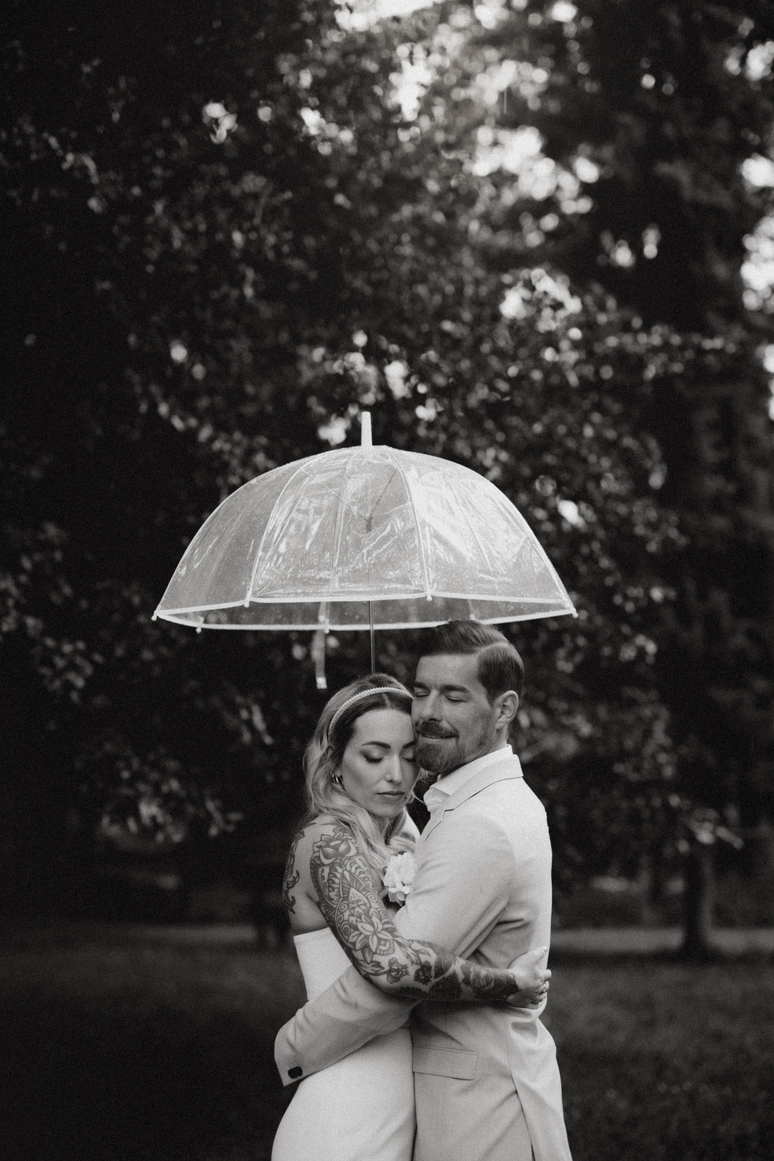 Ein Schwarz-Weiß-Foto von Tasha Ender Photography, Hochzeitsfotografie-Service in Aargau, zeigt ein Paar, das sich eng umarmt unter einem transparenten Regenschirm befindet. Die tätowierte Frau trägt ein Kleid, während der Mann in einem hellen Anzug sie sanft hält, während der Regen sanft um sie herum fällt und ihre Augen geschlossen sind. Bäume bilden einen verschwommenen Hintergrund.