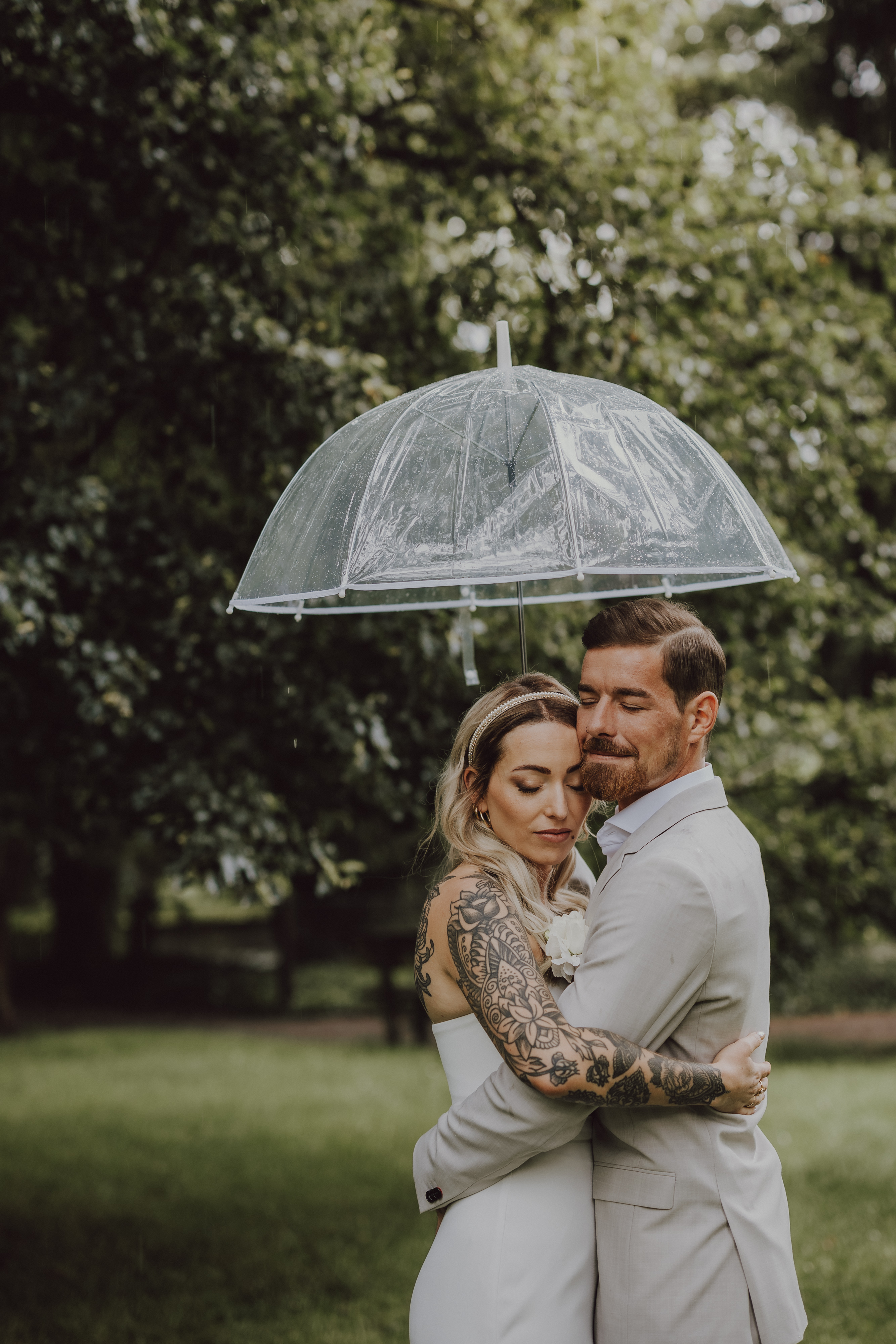 Ein Paar umarmt sich unter einem transparenten Sonnenschirm im Freien während einer romantischen Hochzeitsfotografie-Session von Tasha Ender Photography. Der bärtige Mann trägt einen hellgrauen Anzug, während die Frau, die sichtbare Tattoos auf ihren Armen zeigt, ein weißes Kleid trägt. Saftig grüne Bäume und Gras bilden den Hintergrund dieser bezaubernden Aargauer Szene im Regen.