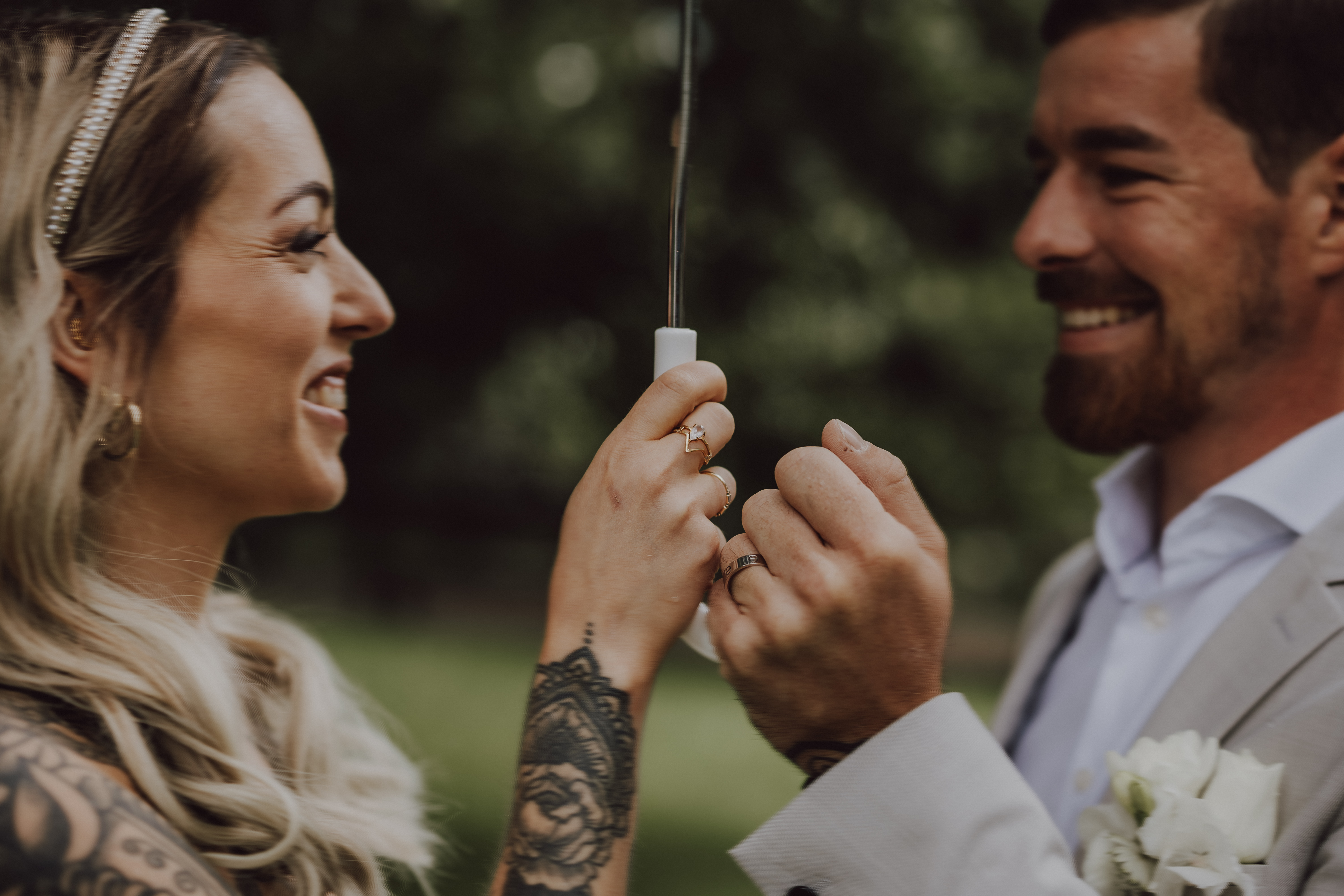 In dieser Hochzeitsfotografie von Tasha Ender Photography steht sich ein Paar lächelnd gegenüber und hält gemeinsam eine Wunderkerze in der Hand. Die Frau, geschmückt mit langen blonden Haaren und einem Stirnband, präsentiert ein Tattoo-Ärmel. Der Mann, der einen hellen Anzug mit kurzen Haaren und Bart trägt, hält einen kleinen Blumenstrauß in der Hand. Ihr Fotoshooting findet vor einer unscharfen Außenkulisse in Regen, Aargau, statt.