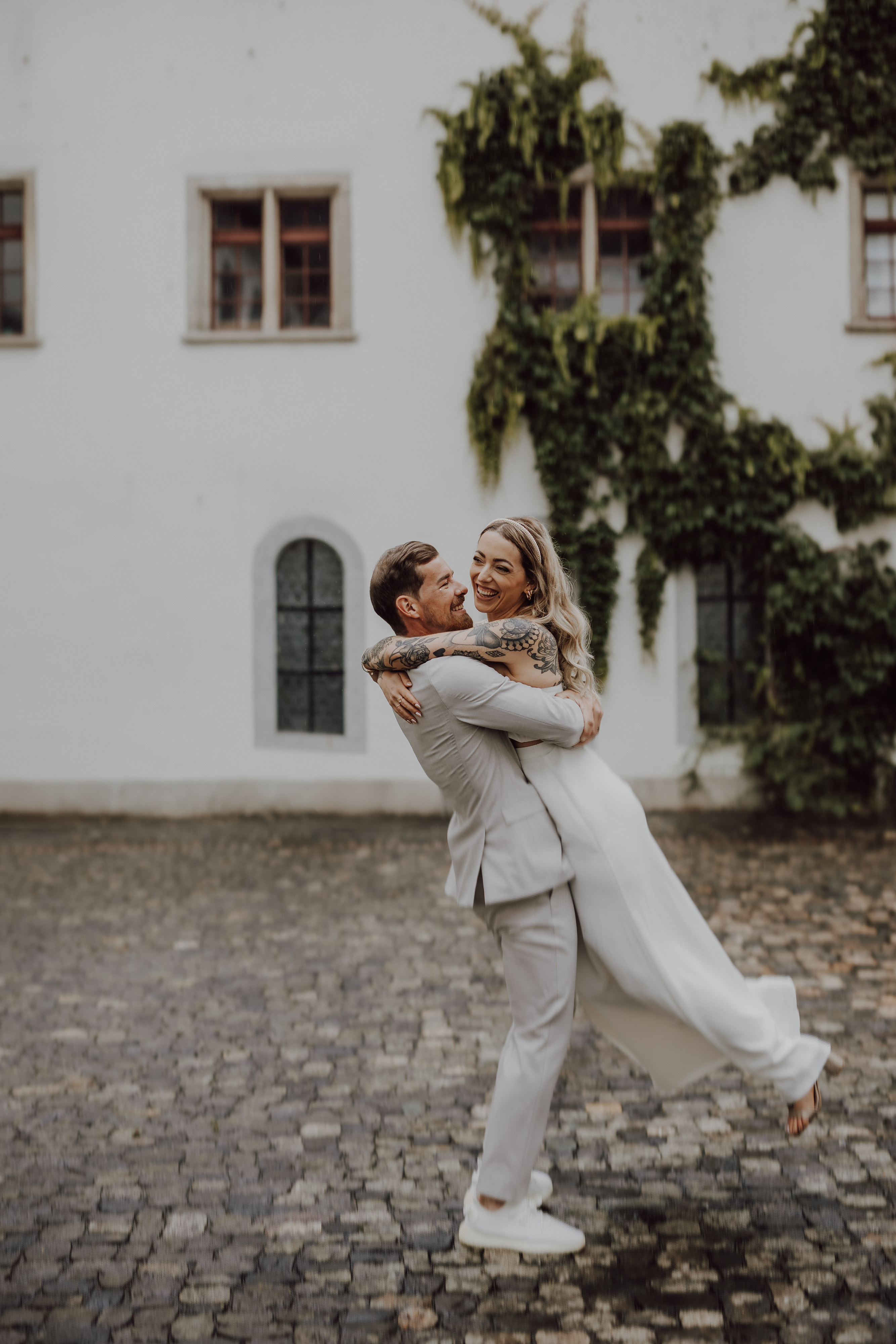Ein Paar in Hochzeitskleidung posiert fröhlich in einem Innenhof. Der Bräutigam, der einen hellen Anzug trägt, hebt die Braut hoch, die ein fließendes weißes Kleid trägt. Sie lächeln und umarmen sich während ihrer Hochzeitsfotografie-Sitzung von Tasha Ender Photography vor einem mit grünen Weinreben bedeckten Gebäude im Hintergrund.