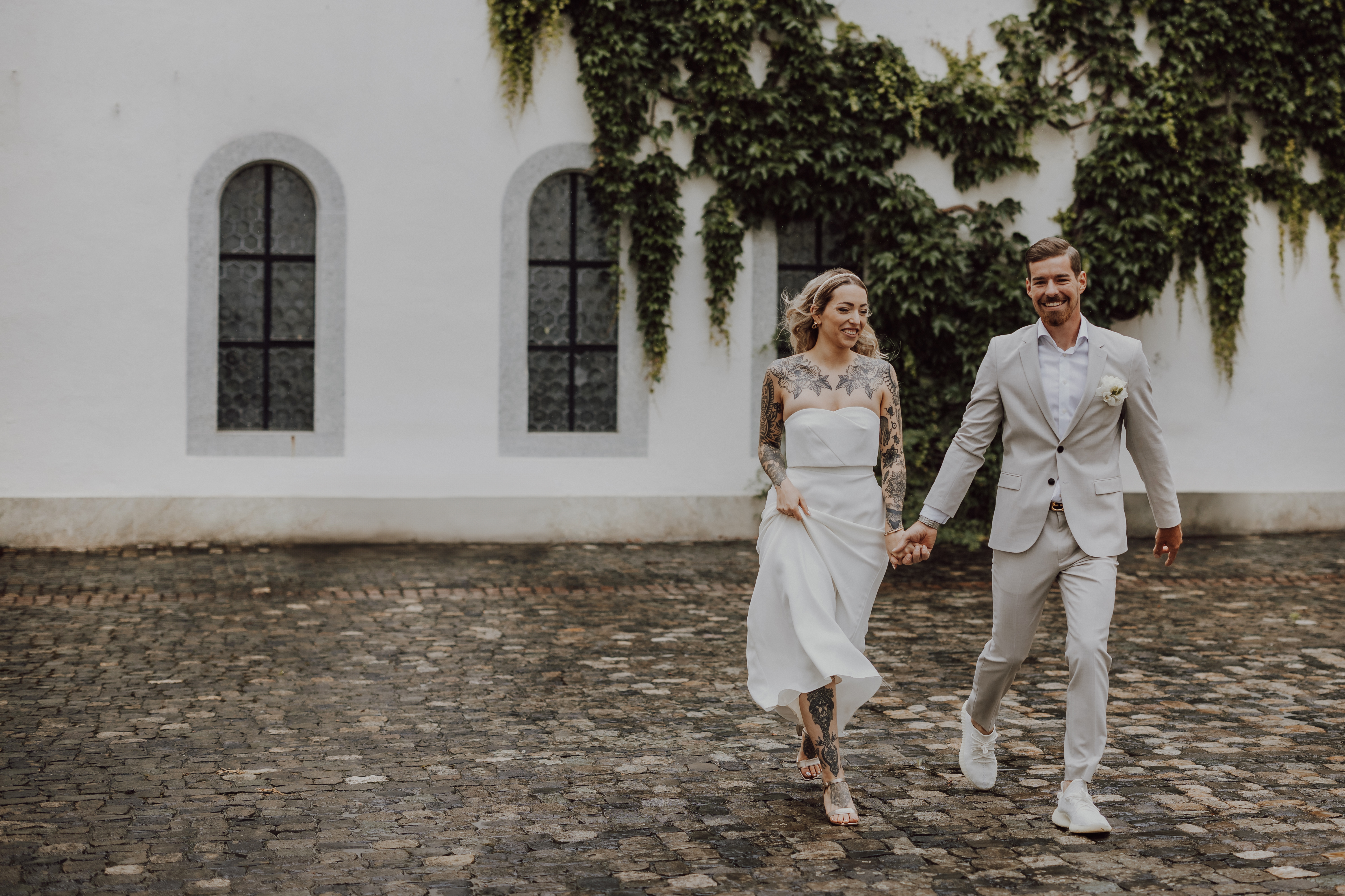 Ein Paar in Hochzeitskleidung geht Hand in Hand. Der Bräutigam, Tasha Ender, trägt einen hellgrauen Anzug und die Braut ein weißes Kleid mit Spitzenärmeln. Beide lächeln und wirken fröhlich. Sie gehen auf einem Kopfsteinpflasterweg mit einem weißen Gebäude und üppigem grünem Efeu im Hintergrund ihrer Hochzeitsszene.