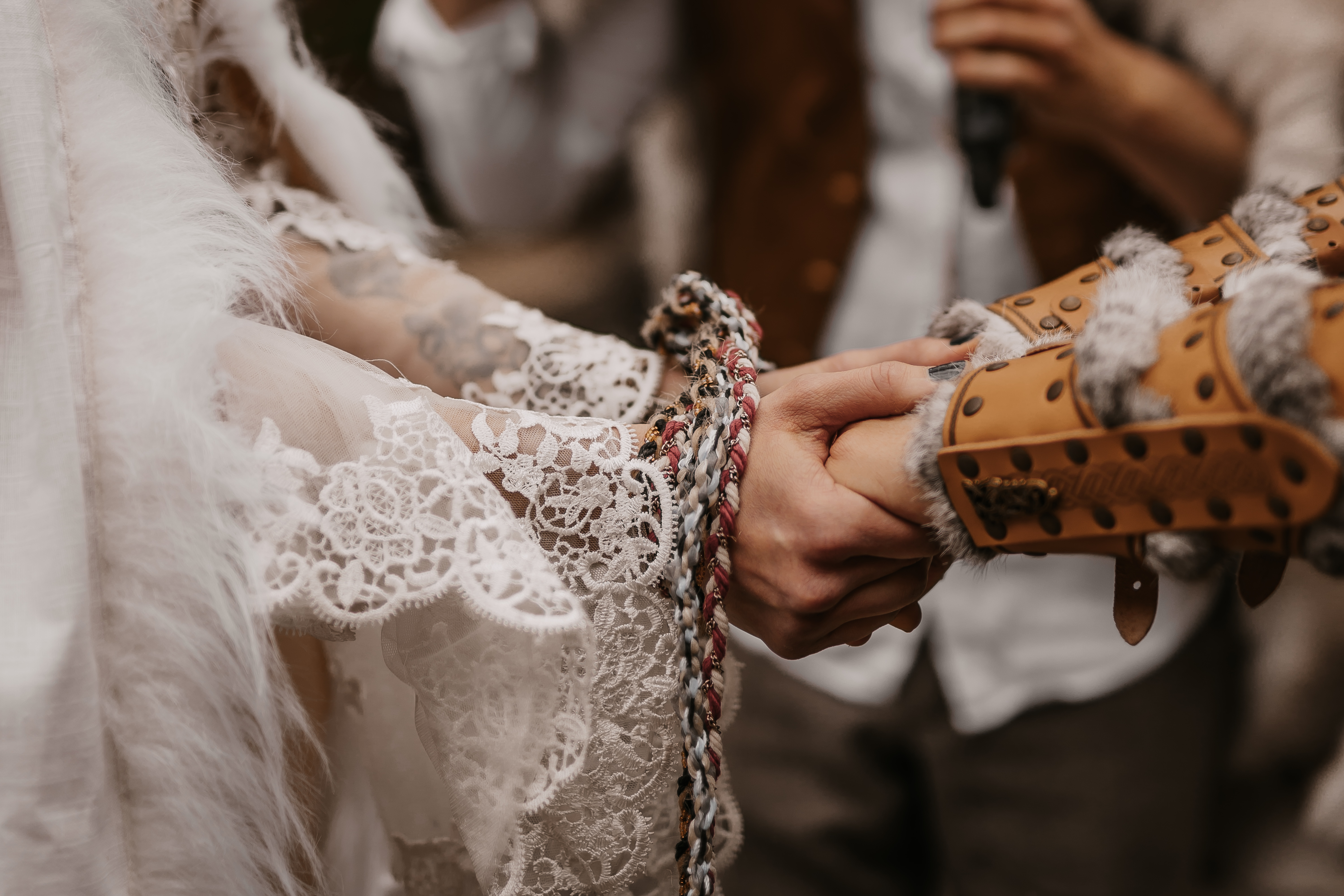 Eine Nahaufnahme der Hände eines Paares, die bei einer Handfasting-Zeremonie zusammengelegt werden, aufgenommen von Tasha Ender Photography. Die Hände sind mit bunten geflochtenen Kordeln aus ökologischen Materialien zusammengebunden, was den einzigartigen Stil der Hochzeitsfotografie widerspiegelt. Eine Person trägt einen Spitzenärmel, während die andere einen mit Pelz und Leder verzierten Armschutz trägt. Im Hintergrund ist ein Geistlicher zu sehen.