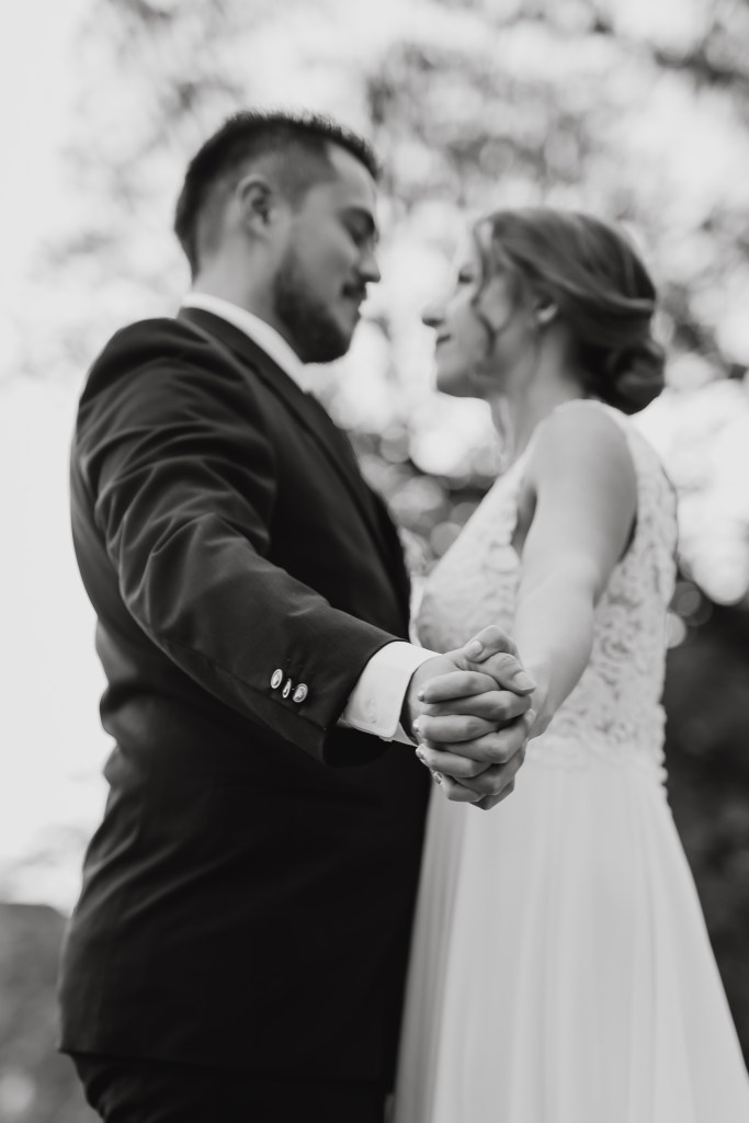 Ein Schwarzweißfoto eines sich umarmenden Paares in formeller Kleidung. Die Braut, Tasha Ender, trägt ein elegantes Kleid und der Bräutigam einen Anzug. Sie halten Händchen und schauen einander in die Augen, was vor einem verschwommenen Naturhintergrund ein Gefühl von Liebe und Intimität vermittelt und ihre Hochzeit wunderschön einfängt.