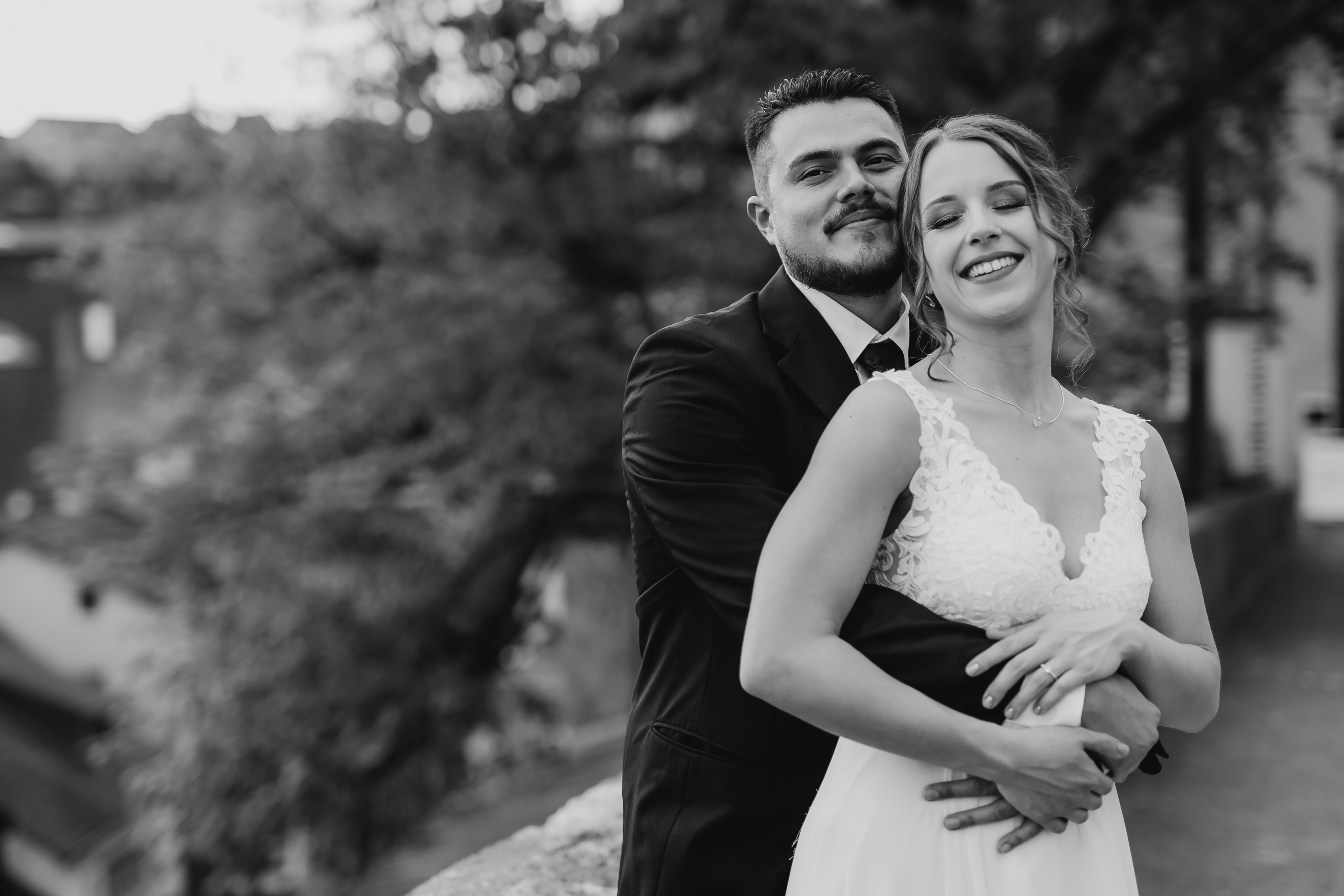 Braut und Bräutigam stehen dicht beieinander und lächeln, während der Bräutigam seine Arme um die Braut legt. Sie tragen einen schwarzen Anzug bzw. ein weißes Hochzeitskleid. Der Hintergrund ist eine verschwommene Außenkulisse mit Bäumen und Gebäuden in Baden. Das Bild, aufgenommen von ihrer Hochzeitsfotografin, ist in Schwarzweiß.