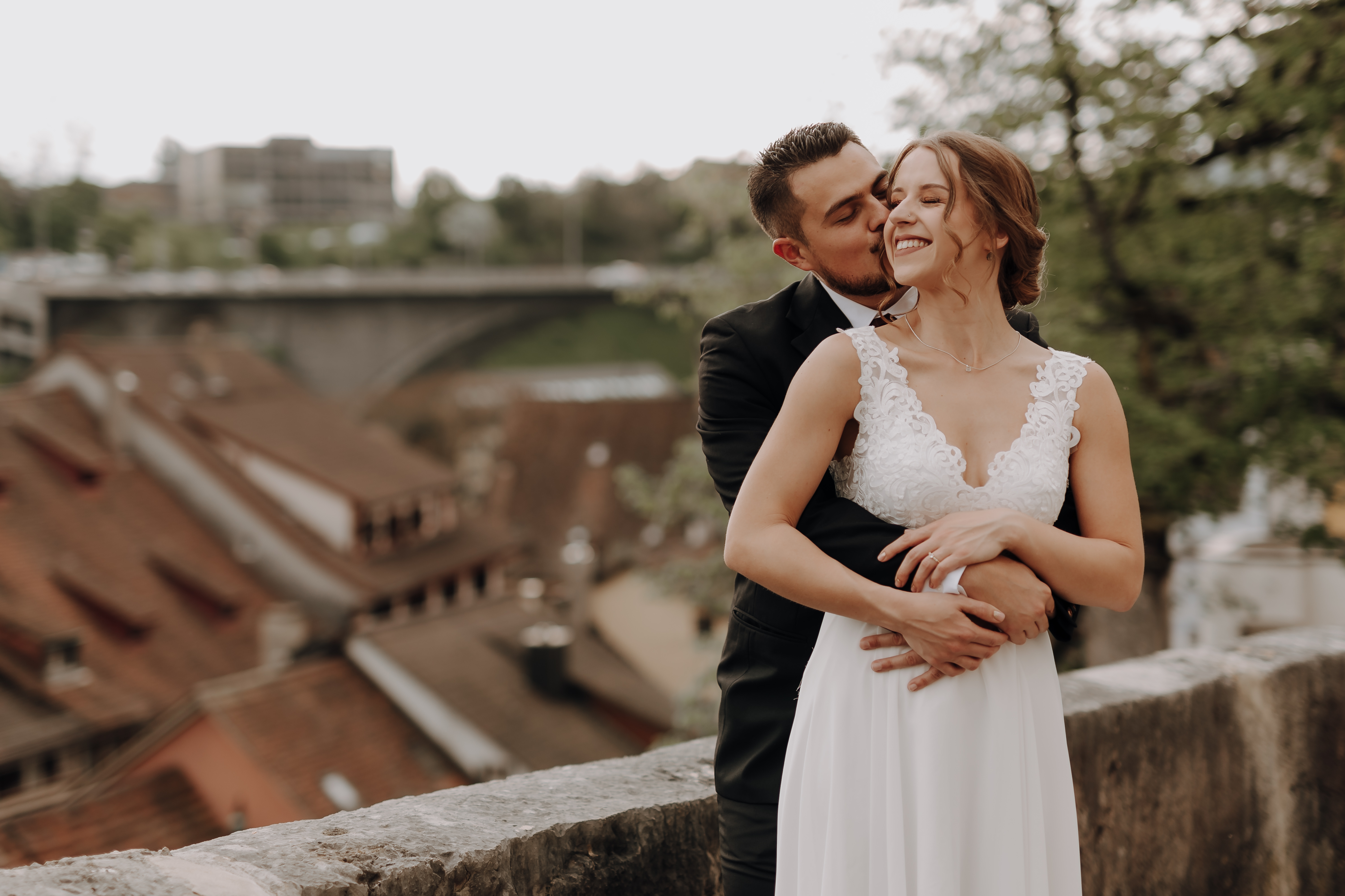 Ein Bräutigam im schwarzen Anzug umarmt eine lächelnde Braut im weißen Spitzenbrautkleid von hinten. Sie stehen auf einem Felsvorsprung mit Dächern und einer Brücke im Hintergrund, perfekt für Hochzeitsfotografie. Im Hintergrund sind Bäume und Gebäude zu sehen.
