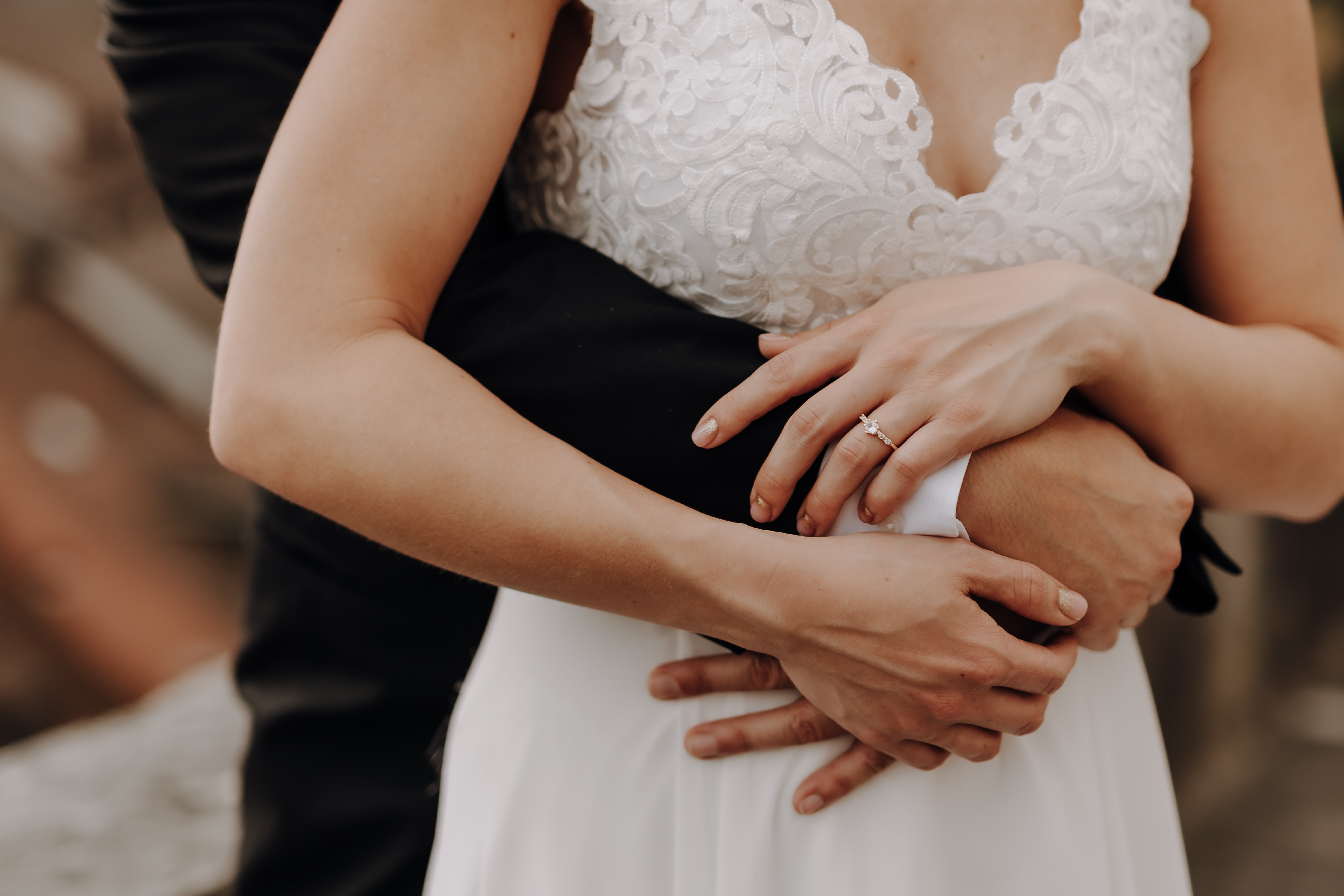 Eine Nahaufnahme einer Braut in einem weißen Spitzenhochzeitskleid, die von hinten von einer Person in einem schwarzen Anzug umarmt wird. Ihr Verlobungsring ist sichtbar, während sich ihre Hände sanft um ihre Taille legen. Perfekt eingefangen von Hochzeitsfotografin Tasha Ender während der Ziviltrauung.