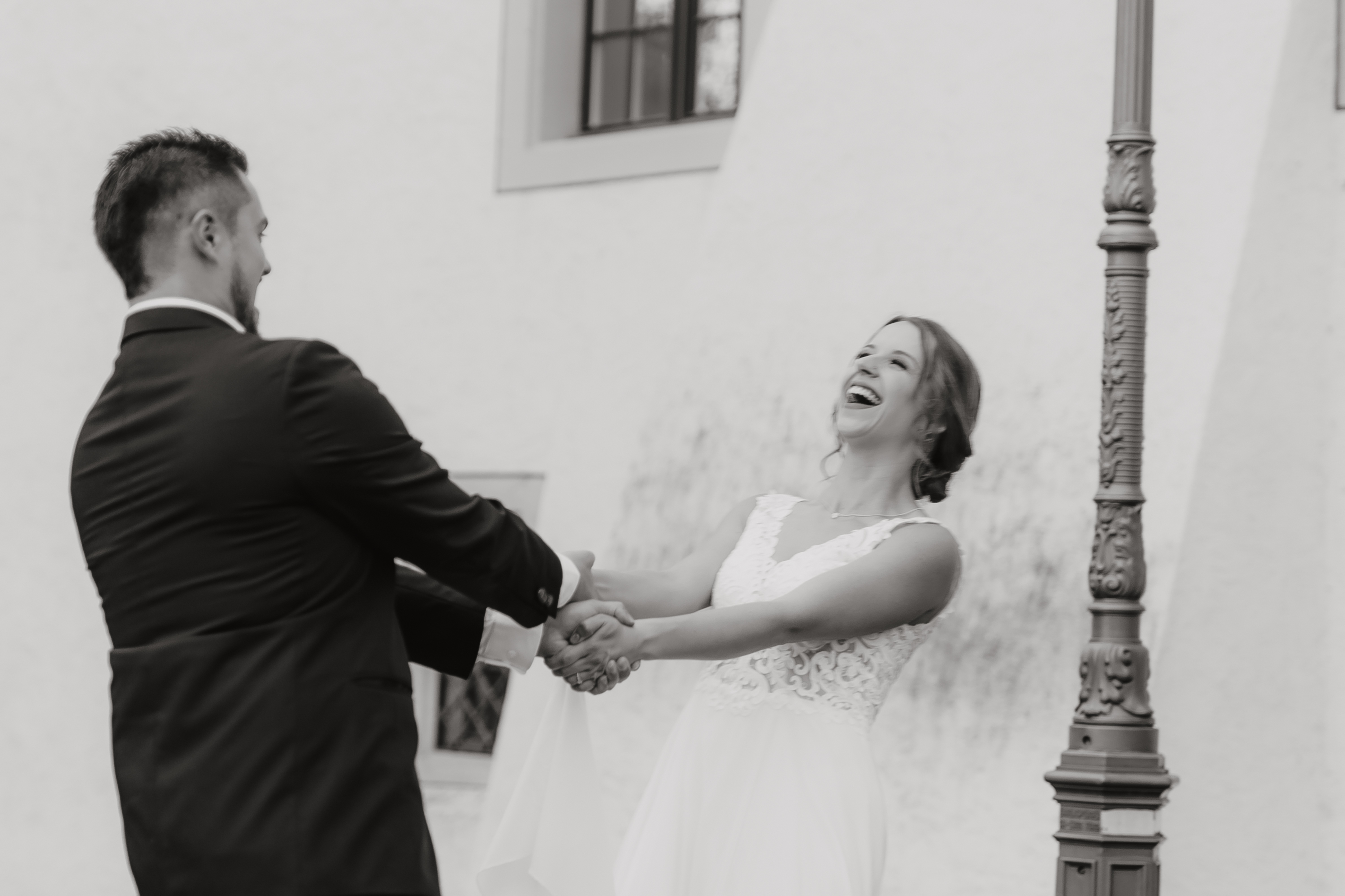 Schwarzweißfoto einer fröhlichen Braut und eines Bräutigams, die Händchen halten und lachen. Die Braut trägt ein weißes Hochzeitskleid mit Spitzendetails und der Bräutigam einen Anzug. Sie scheinen während ihrer Ziviltrauung vor einem eleganten Gebäude mit einem Laternenpfahl in der Nähe zu tanzen oder zu wirbeln.