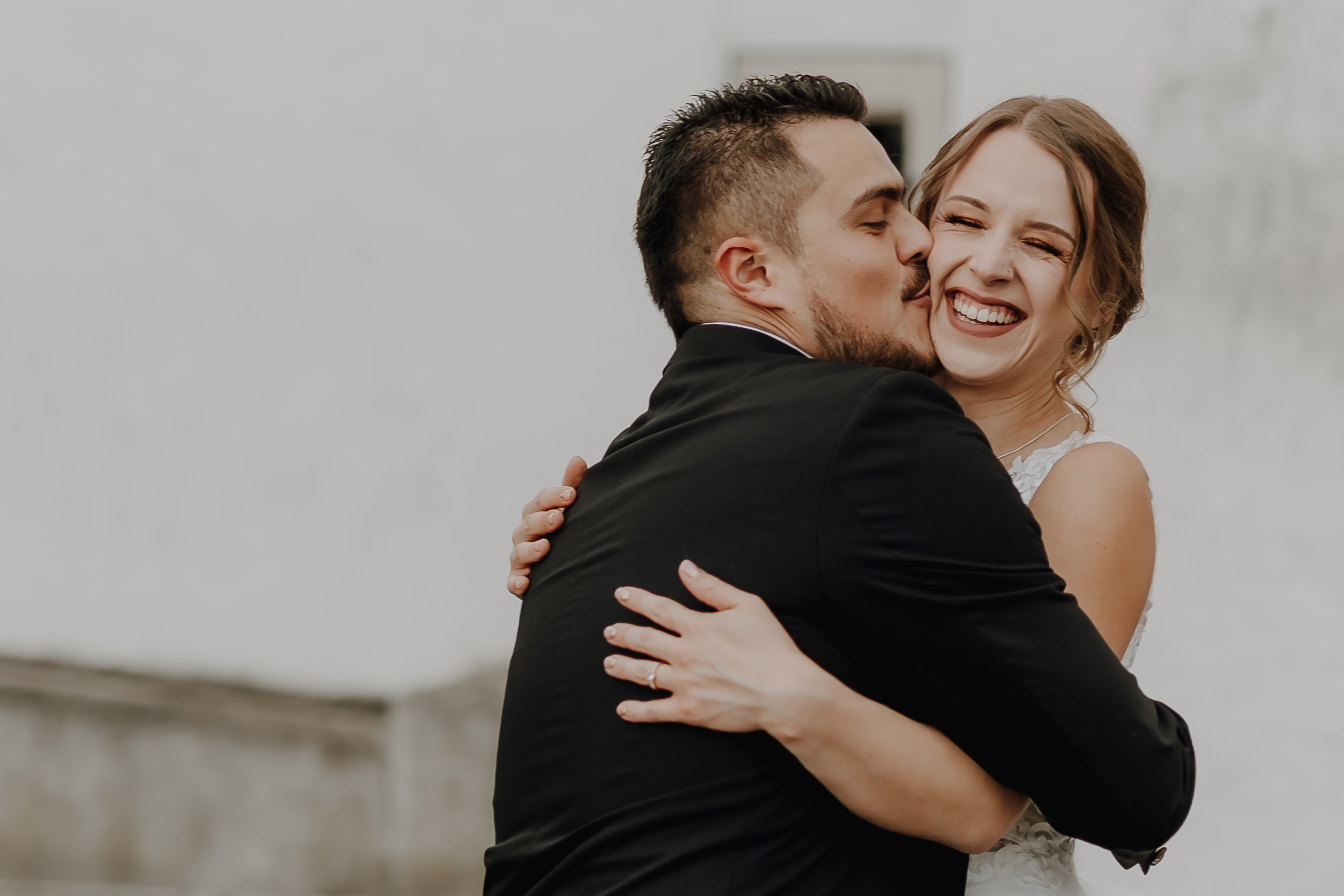 Braut und Bräutigam umarmen sich freudig an ihrem Hochzeitstag. Der Bräutigam, in einen schwarzen Anzug gekleidet, küsst die Braut auf die Wange. Die Braut, in einem weißen Brautkleid, lächelt strahlend mit geschlossenen Augen vor einem verschwommenen, hellen Hintergrund während ihrer Hochzeitsfotografie-Sitzung von Tasha Ender Photography für ihr Zivilhochzeits-Fotoshooting in Baden.