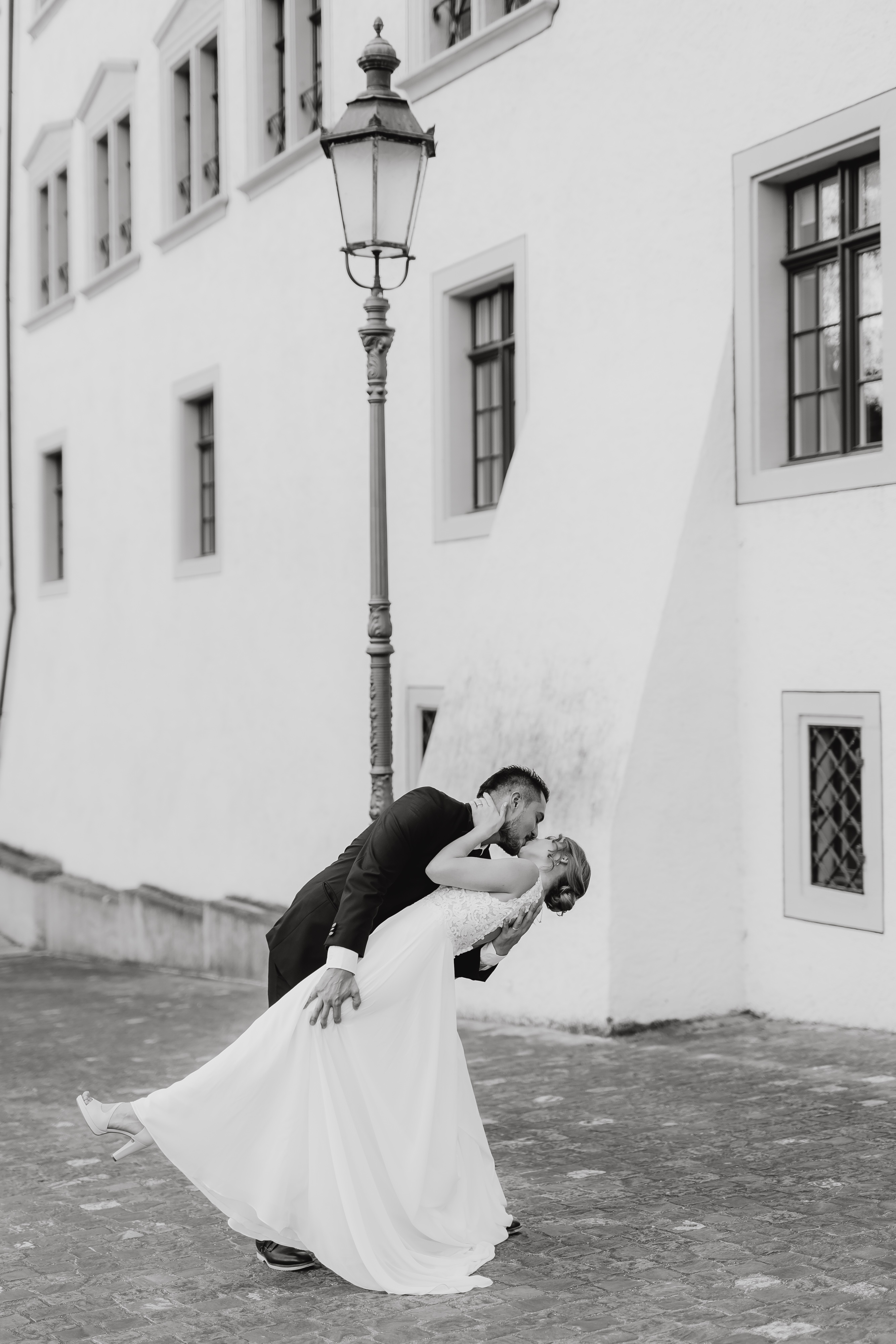 Ein frisch vermähltes Paar tanzt vor einem Gebäude mit mehreren Fenstern. Der Bräutigam, der einen schwarzen Anzug trägt, taucht freudig die Braut in ein fließendes weißes Kleid. Sie sind von Kopfsteinpflaster umgeben und eine klassische Straßenlaterne trägt zur romantischen Atmosphäre ihrer Zivilhochzeit in Baden bei. Dieser magische Moment wurde vom Hochzeitsfotografie-Service von Tasha Ender Photography wunderschön festgehalten.