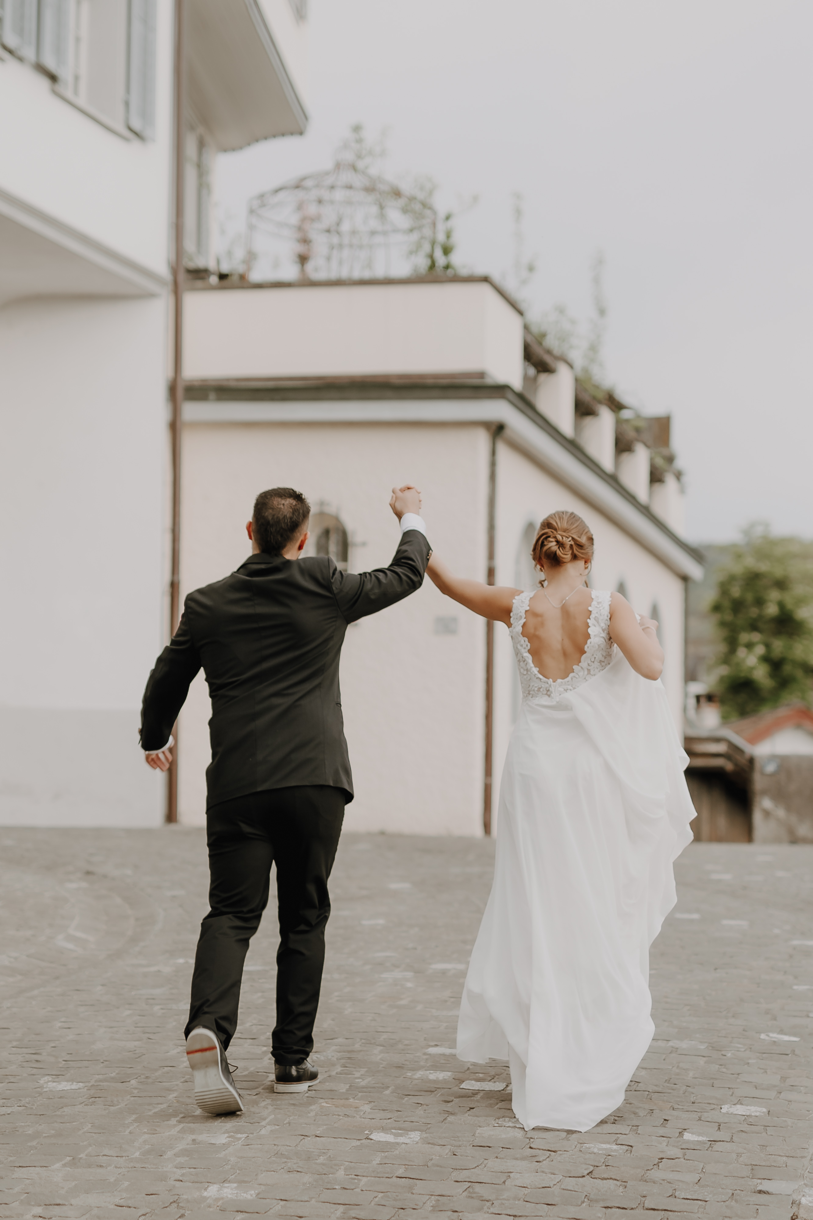 Ein Paar ist von hinten zu sehen, wie es Hand in Hand geht. Der Mann trägt einen schwarzen Anzug und die Frau ein weißes Hochzeitskleid. Sie scheinen auf einem Kopfsteinpflasterweg neben einem Gebäude mit weißen Wänden und Blumenkästen voller Pflanzen zu stehen, möglicherweise während ihrer Zivilhochzeit im Aargau, wunderschön eingefangen von Hochzeitsfotografie von Tasha Ender Photography.