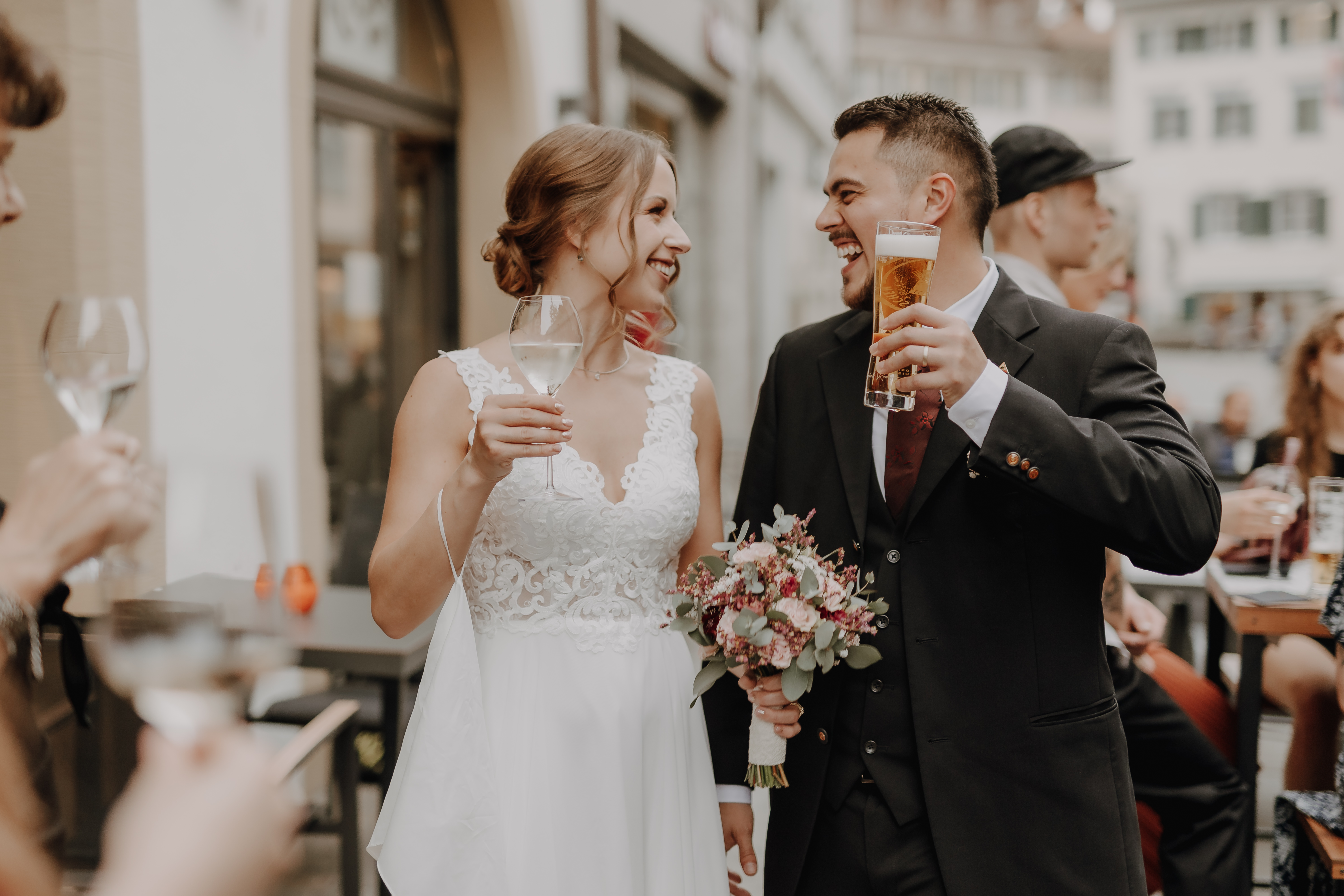 Tasha Ender Photography hat eine fröhliche Zivilhochzeit festgehalten, bei der Braut und Bräutigam im Hochzeitskleid draußen stehen, lächeln und Getränke in den Händen halten. Die Braut glänzt in einem weißen Spitzenkleid mit einem Blumenstrauß in der Hand, während der Bräutigam sie in einem schwarzen Anzug perfekt ergänzt und sein Bier genießt. Sie feiern ihre Liebe in Baden und sind von Gästen umgeben, die ihre Gläser in ausgelassenem Jubel erheben.
