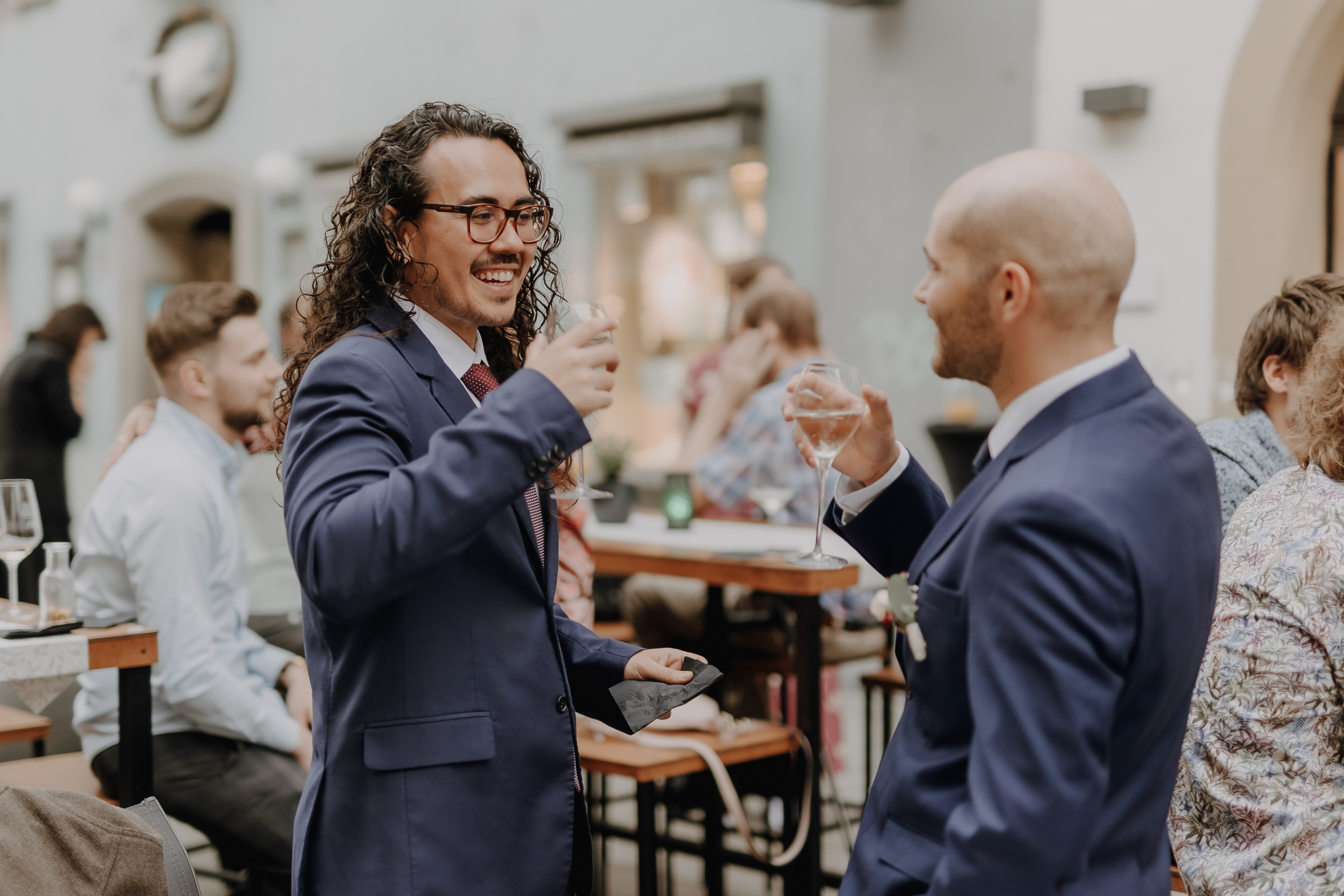 Zwei Männer in Anzügen lächeln und stoßen bei einer gesellschaftlichen Veranstaltung in Baden an. Einer hat langes lockiges Haar und eine Brille, während der andere kahl ist. Sie stehen an einem Stehtisch mit weiteren Personen und einem sanft fokussierten Hintergrund aus Tischen und Gebäudeinnenraum. Diese Versammlung scheint Teil einer Zivilhochzeitsfeier in Aargau zu sein, wunderschön eingefangen vom Hochzeitsfotografie-Service von Tasha Ender Photography.