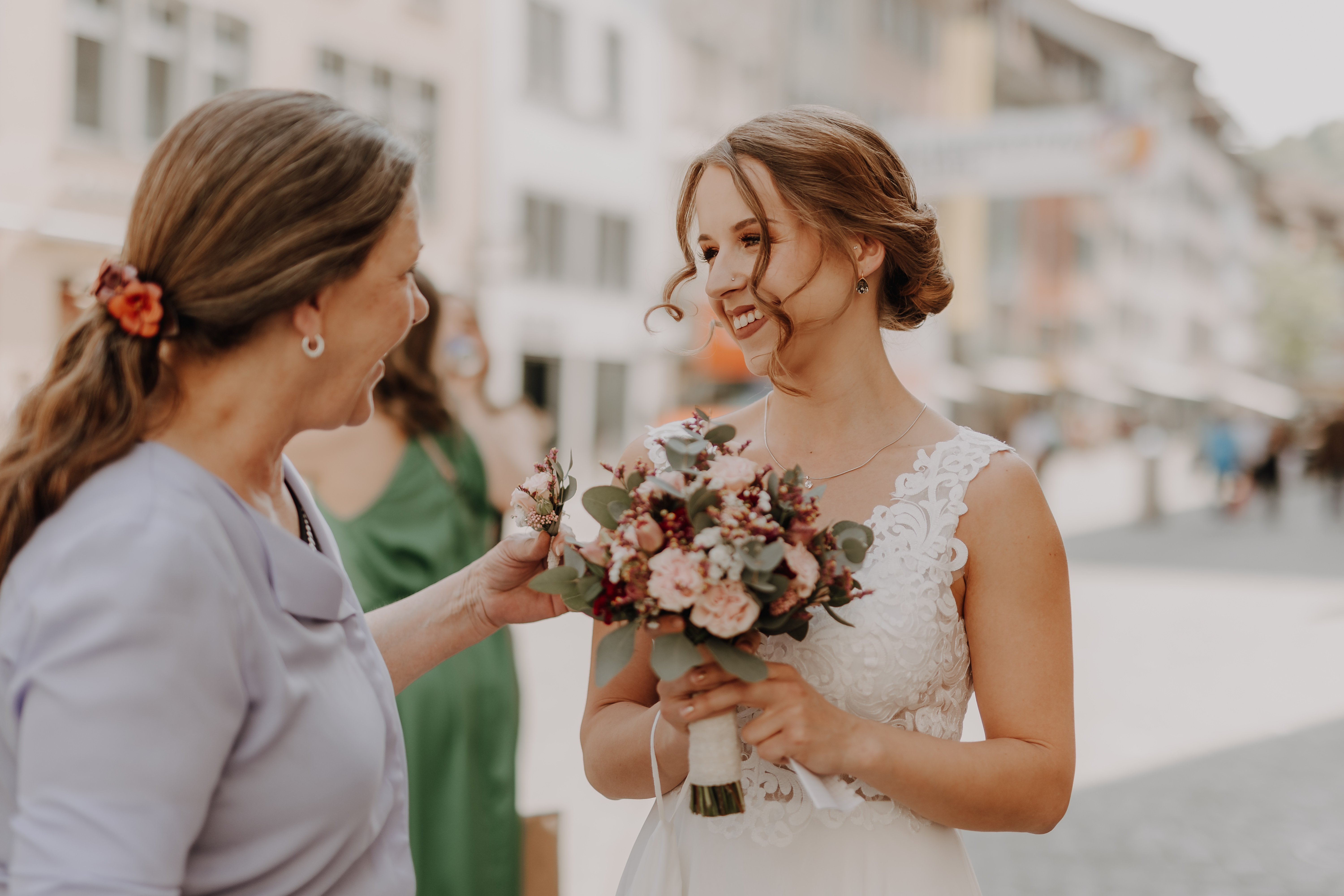 Eine Braut in einem weißen Spitzenhochzeitskleid hält einen Blumenstrauß und lächelt eine andere Frau mit einem lila Oberteil und einer Blume im Haar auf einer Stadtstraße während ihrer Zivilhochzeit an. Aufgenommen von Tasha Ender Photographys Hochzeitsfotografie wirken die beiden glücklich und in ein Gespräch vertieft, mit verschwommenen Gebäuden und anderen Menschen im Hintergrund.