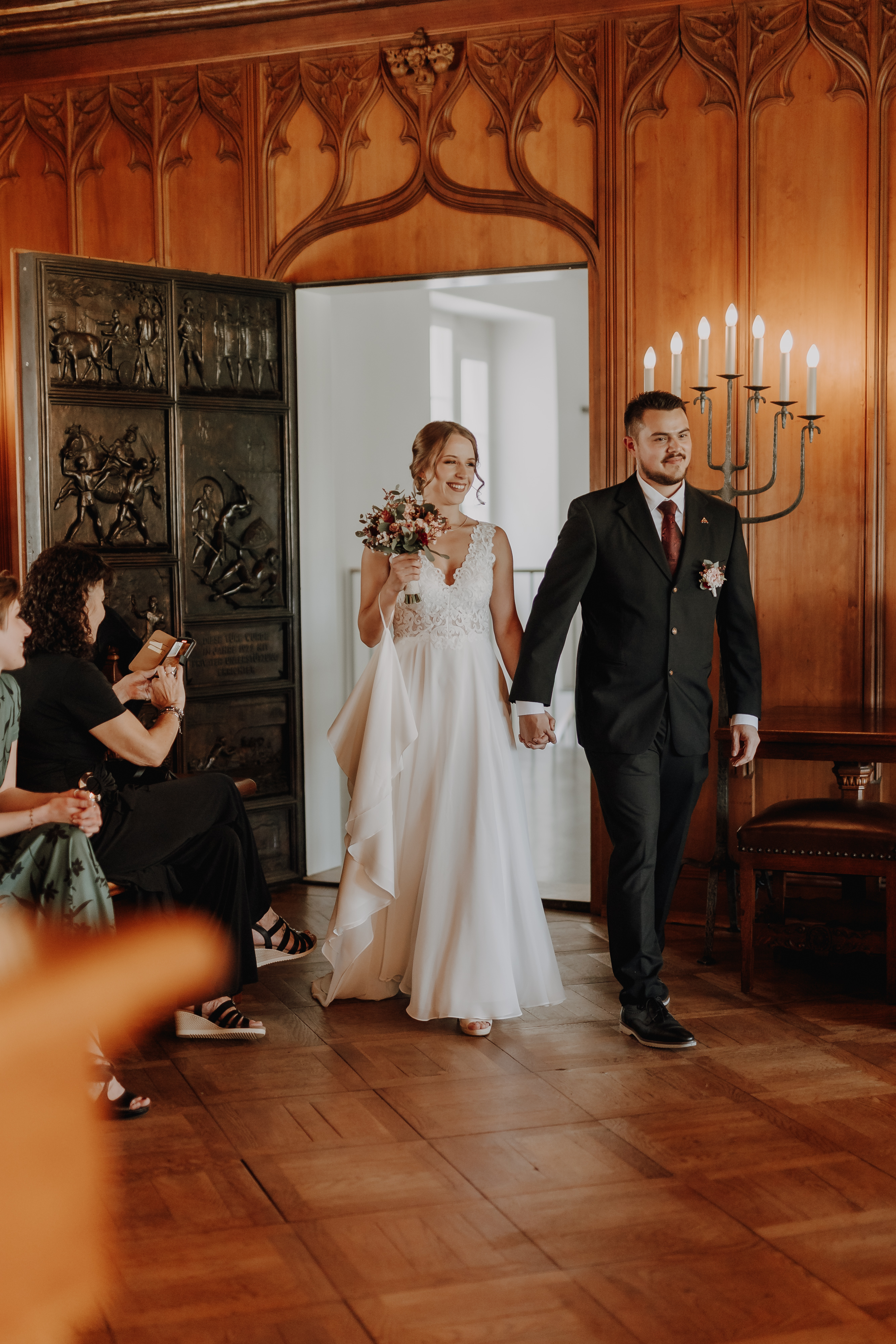 Braut und Bräutigam halten sich an den Händen, als sie einen warm beleuchteten Raum mit Holzvertäfelung und einem Kerzenleuchter betreten, der perfekt für ihre Ziviltrauung ist. Die Braut, in ein weißes Kleid gekleidet und mit einem Blumenstrauß in der Hand, steht neben dem Bräutigam in seinem dunklen Anzug. Gäste, die an der Seite sitzen, schauen aufmerksam zu und halten diesen besonderen Moment mit Hochzeitsfotografie von Tasha Ender Photography fest.