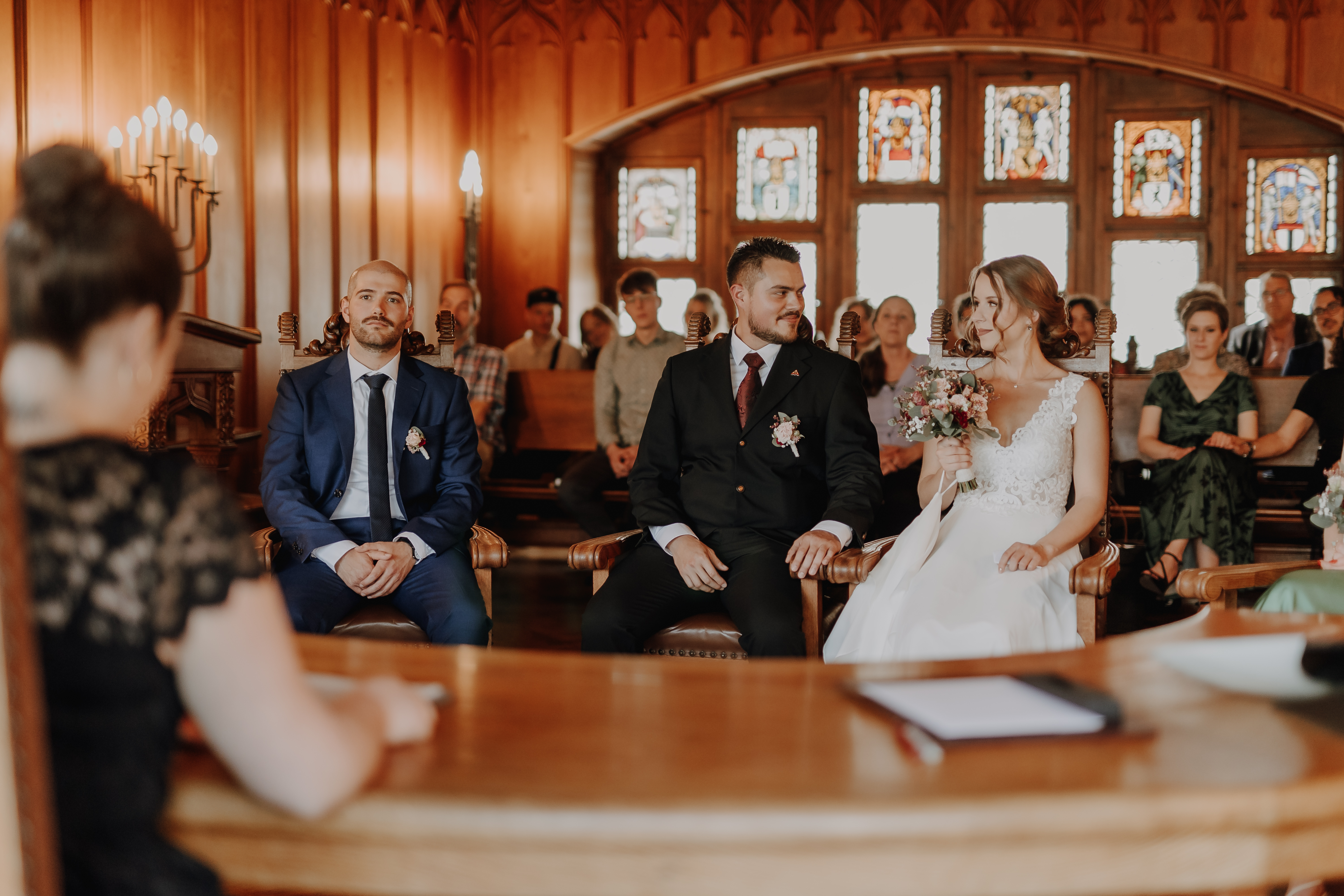 Eine Hochzeitszeremonie findet in einem dekorativen Holzraum mit Buntglasfenstern statt. Die Braut in einem weißen Kleid hält Blumen und sitzt neben dem Bräutigam in einem schwarzen Anzug und einem weiteren Mann in einem blauen Anzug. Die Gäste beobachten von den Kirchenbänken aus, wie ein Geistlicher an seinem Schreibtisch die Zivilhochzeit überwacht, perfekt eingefangen von Hochzeitsfotografie von Tasha Ender Photography.